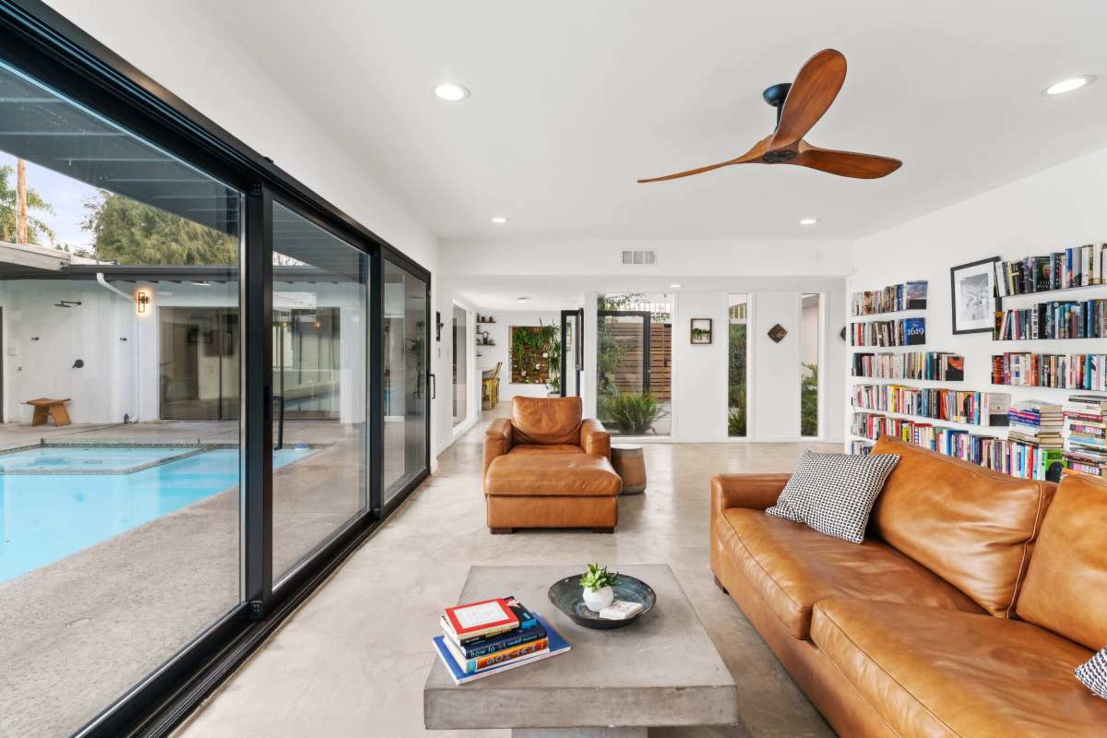 A modern living room features a leather sofa, a coffee table, and large glass doors that open to a pool area.