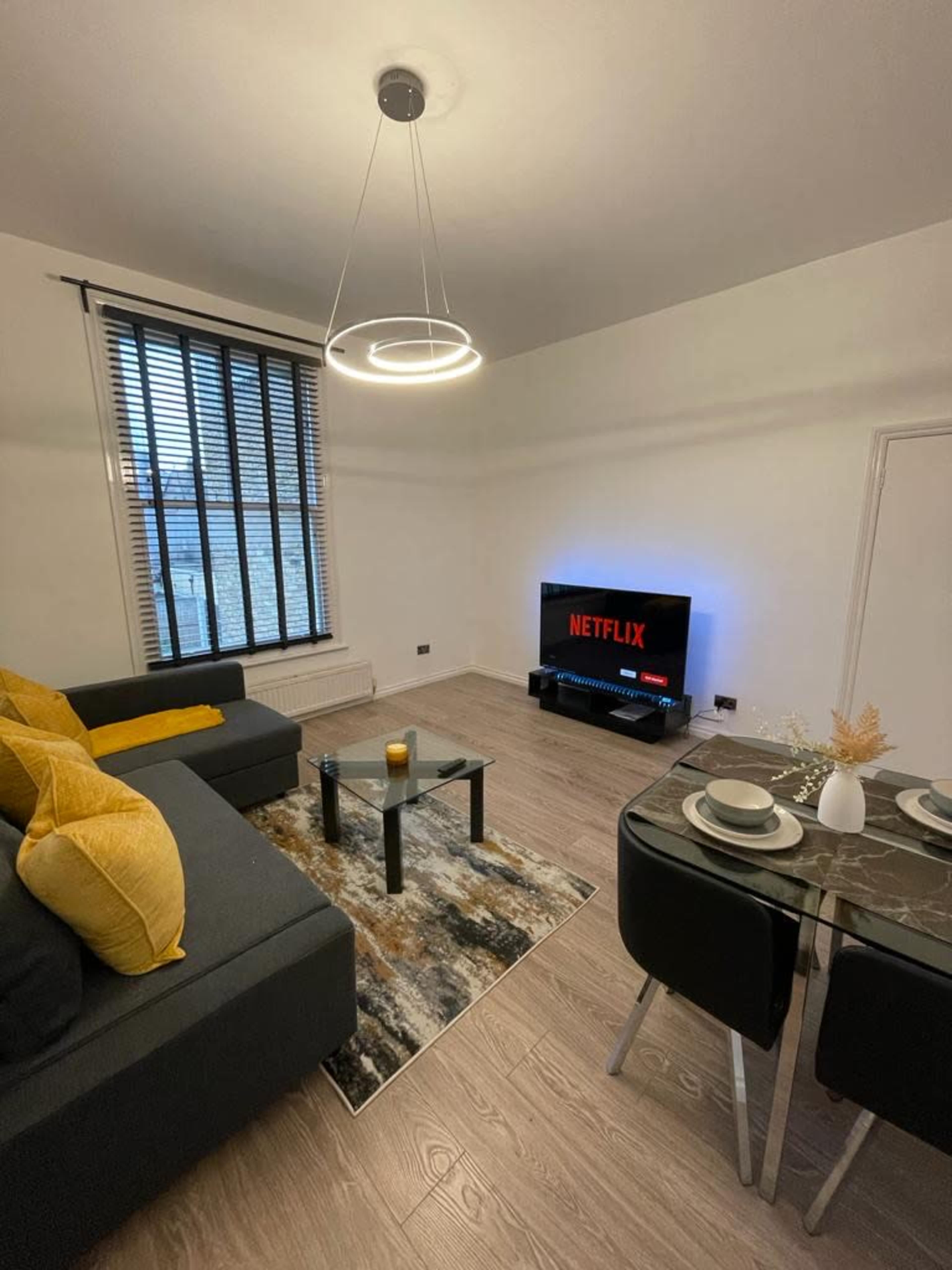 A modern living room featuring a gray sofa with yellow cushions, a round dining table set for two, and a television displaying the Netflix logo.