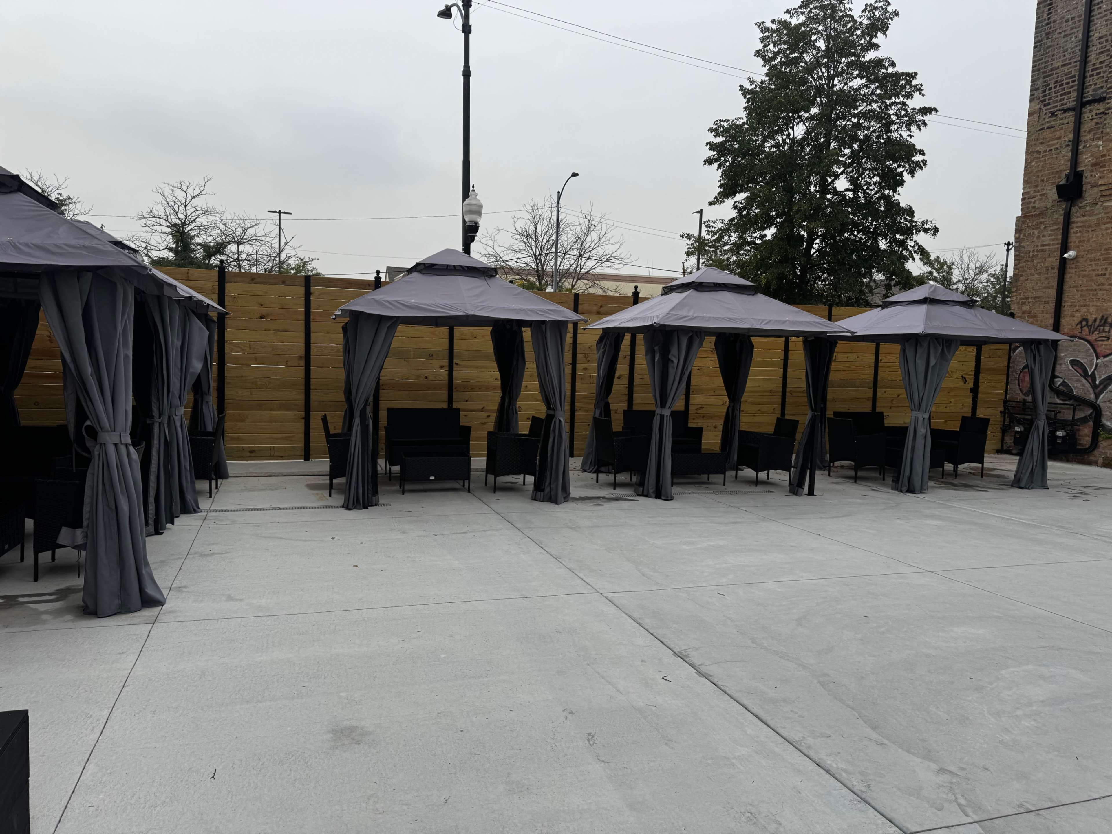 The image shows a series of covered outdoor seating areas with gray canopies, arranged neatly on a concrete surface, and surrounded by a wooden fence.