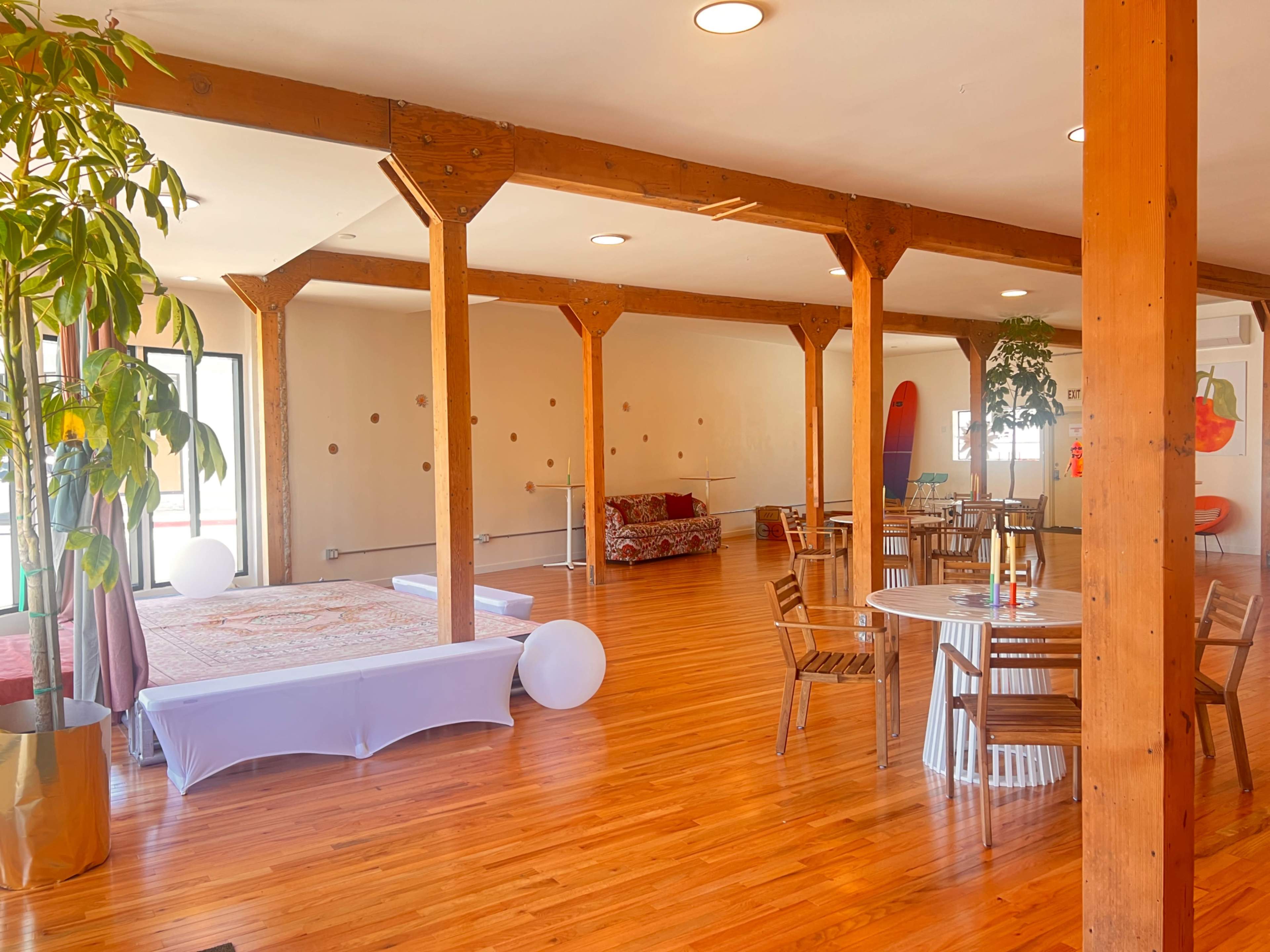 The image shows a spacious interior with wooden pillars, hardwood floors, and various seating arrangements including tables, chairs, and a couch.