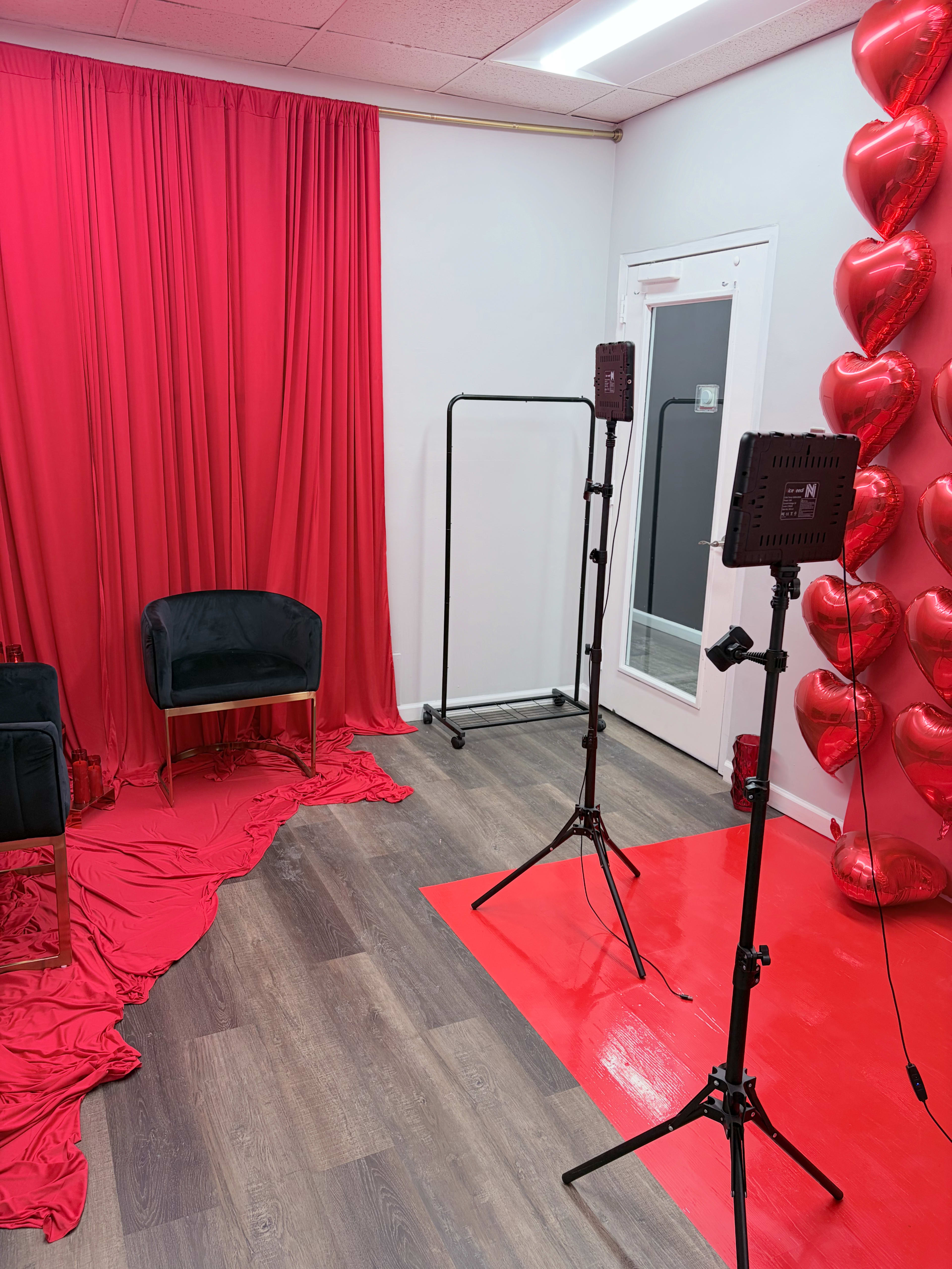 The image shows a small studio space decorated with red curtains, heart-shaped balloons, and a black chair, along with lighting equipment.