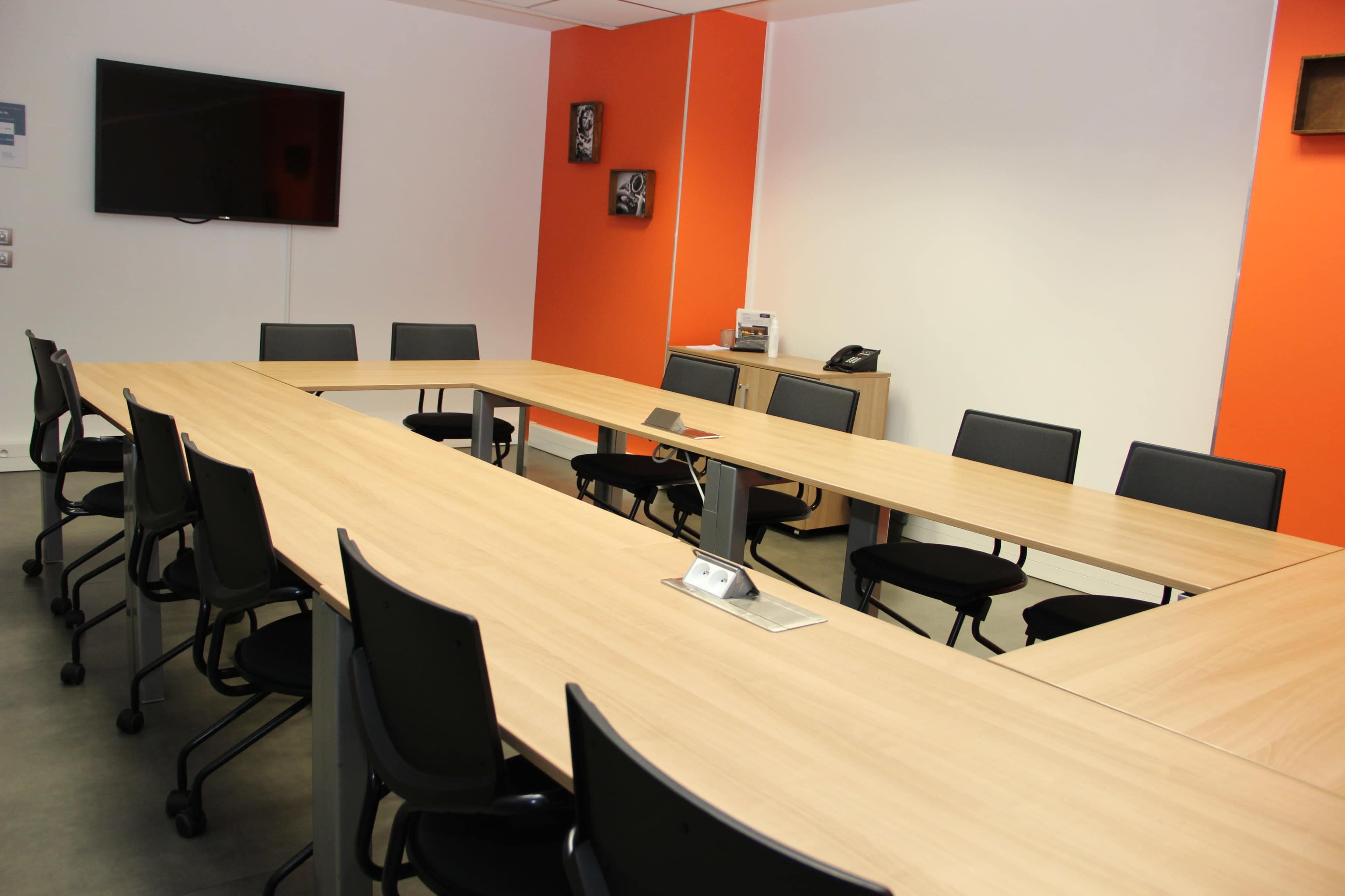 A large conference room features a rectangular table surrounded by black chairs, with an orange accent wall and a television mounted on the white wall.