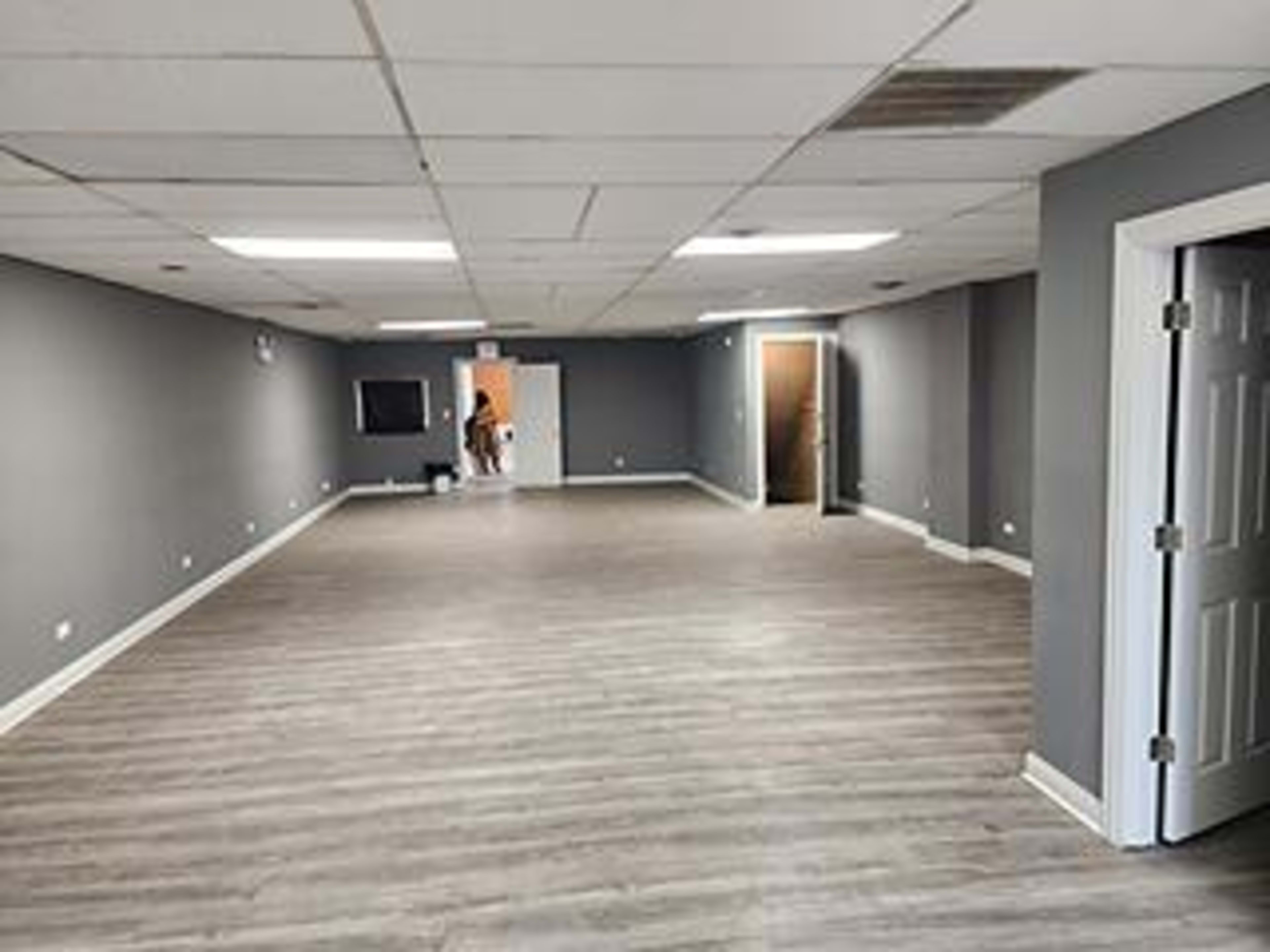 The image shows an empty, spacious room with gray walls, light wood flooring, and a door leading to another area.