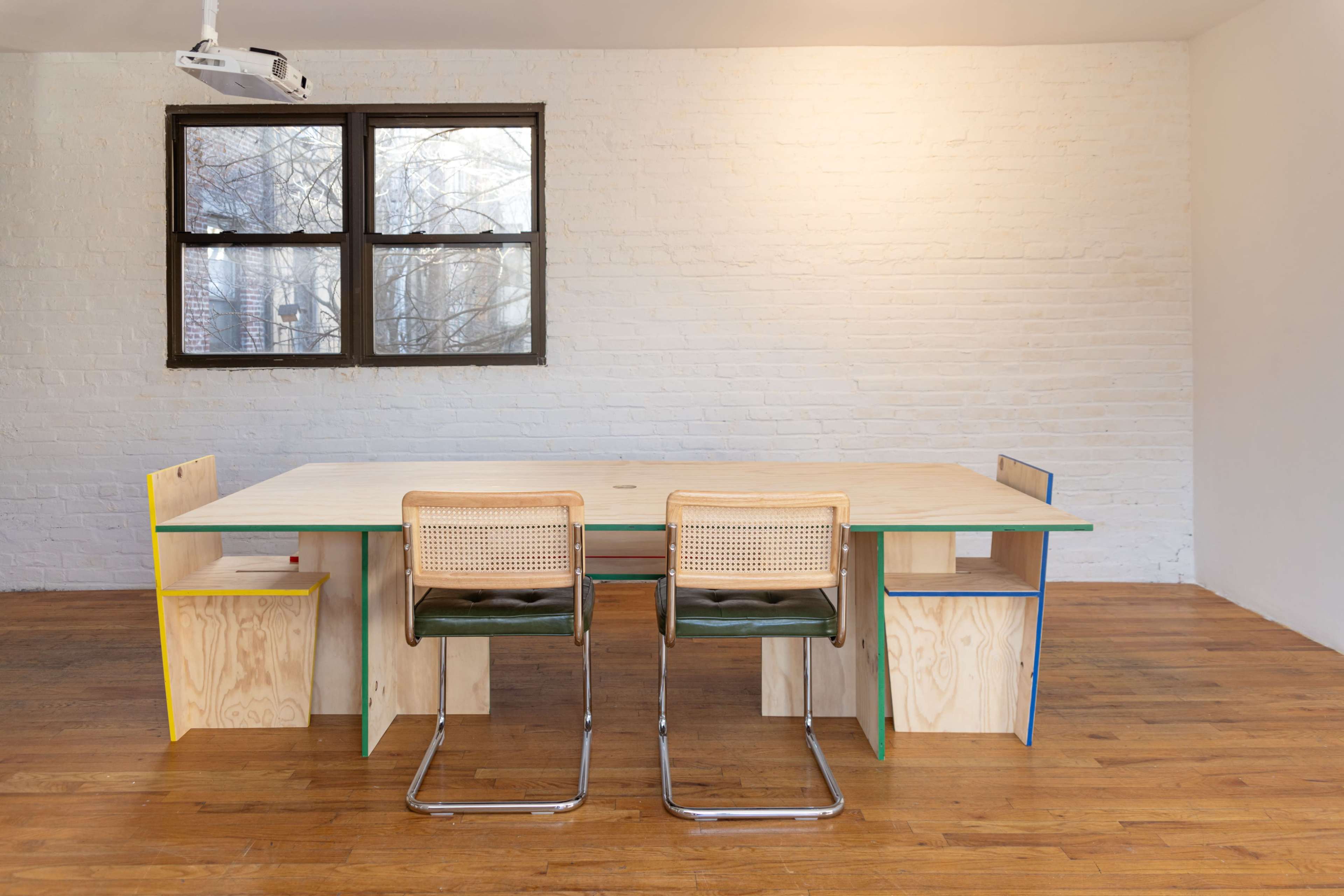 A large, minimalist wooden table with two chairs, positioned in a bright room with a wooden floor and a large window.