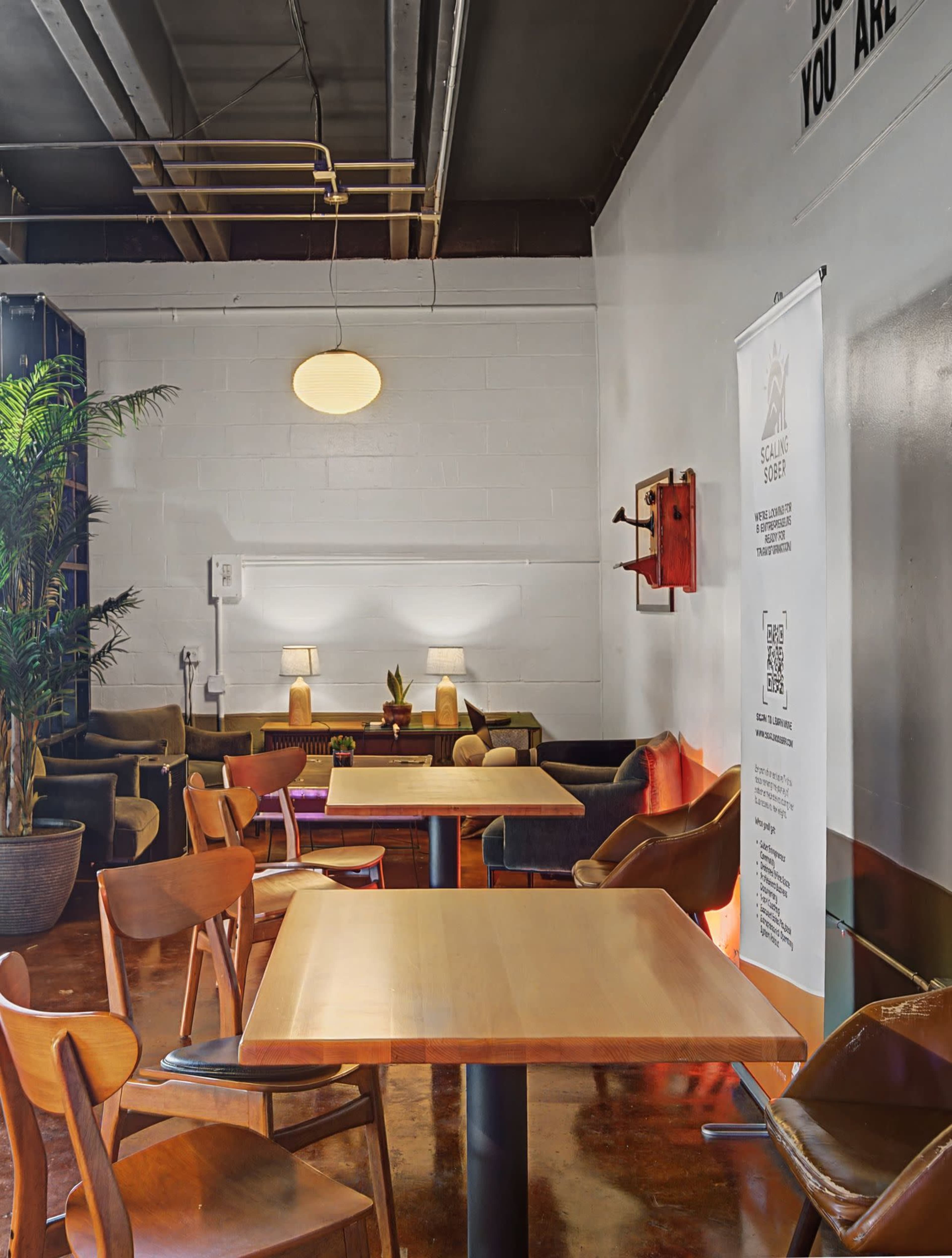 The image shows a cozy café interior with wooden tables, chairs, and warm lighting, complemented by plants and decorative elements on the walls.
