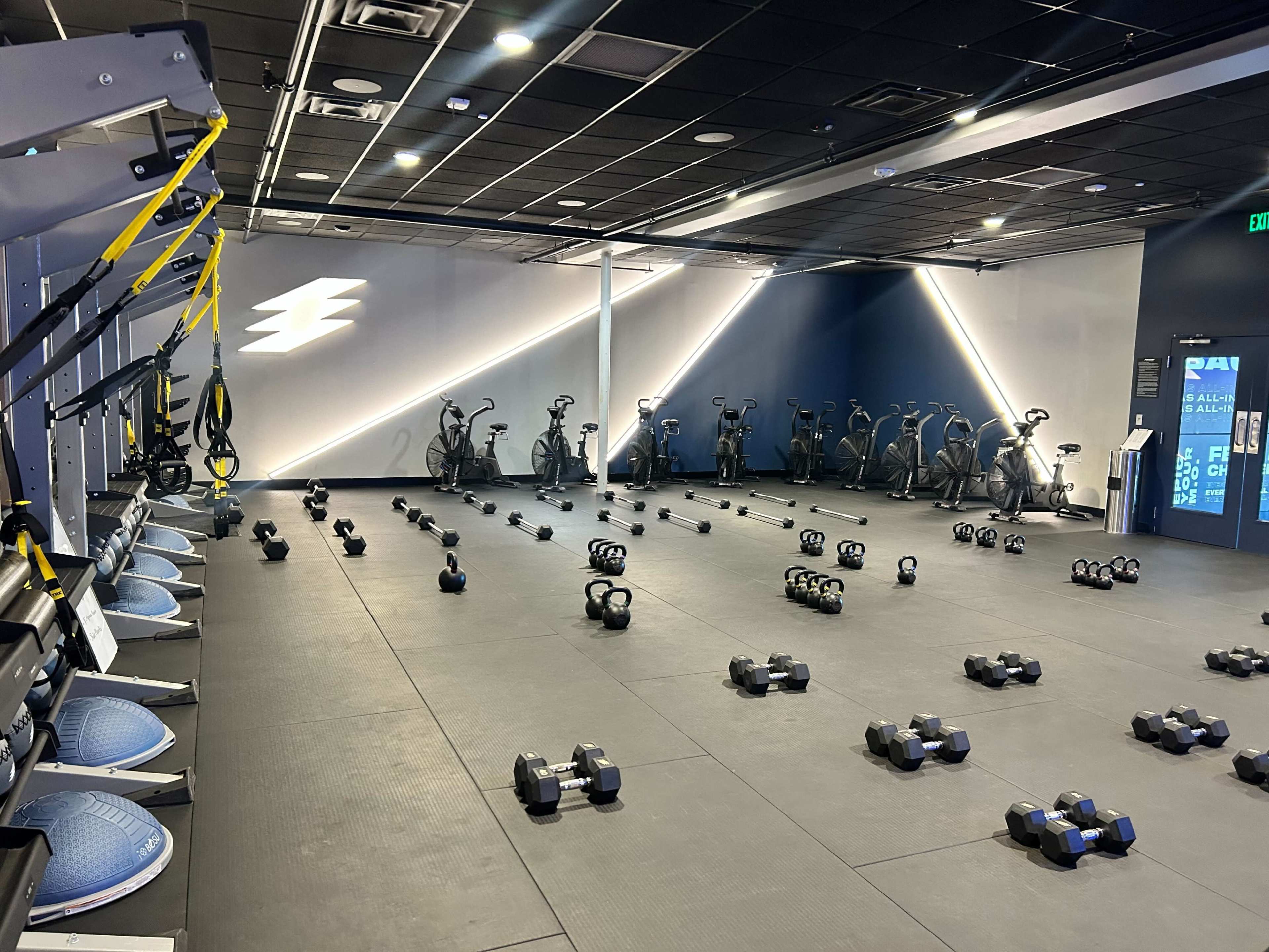 The image shows a modern gym interior with exercise equipment, including rows of dumbbells, stationary bikes, and suspension training straps arranged across a black rubber floor.