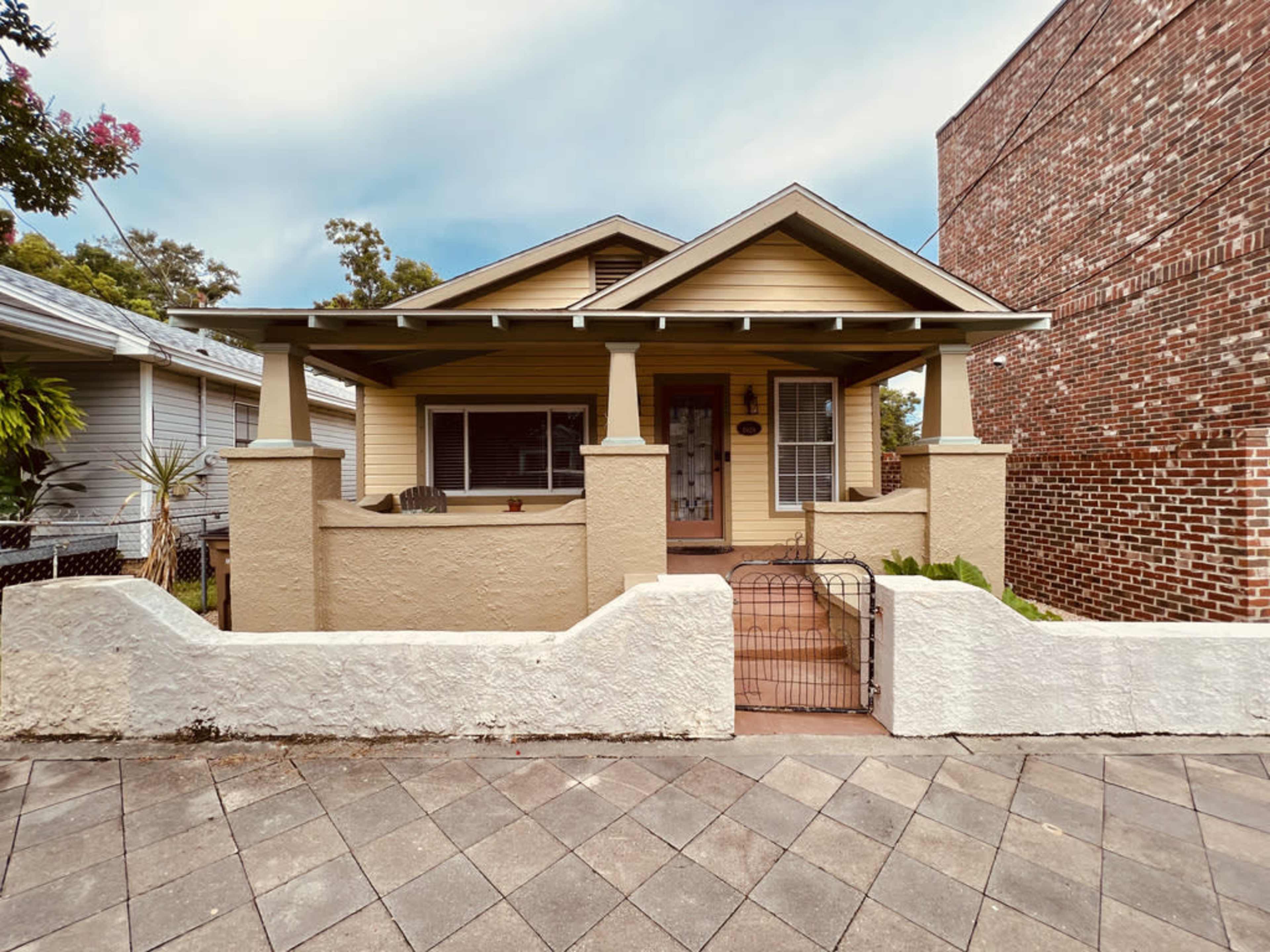 A single-story yellow house with a front porch, flanked by two other houses, and features a low white wall and a brick building in the background.