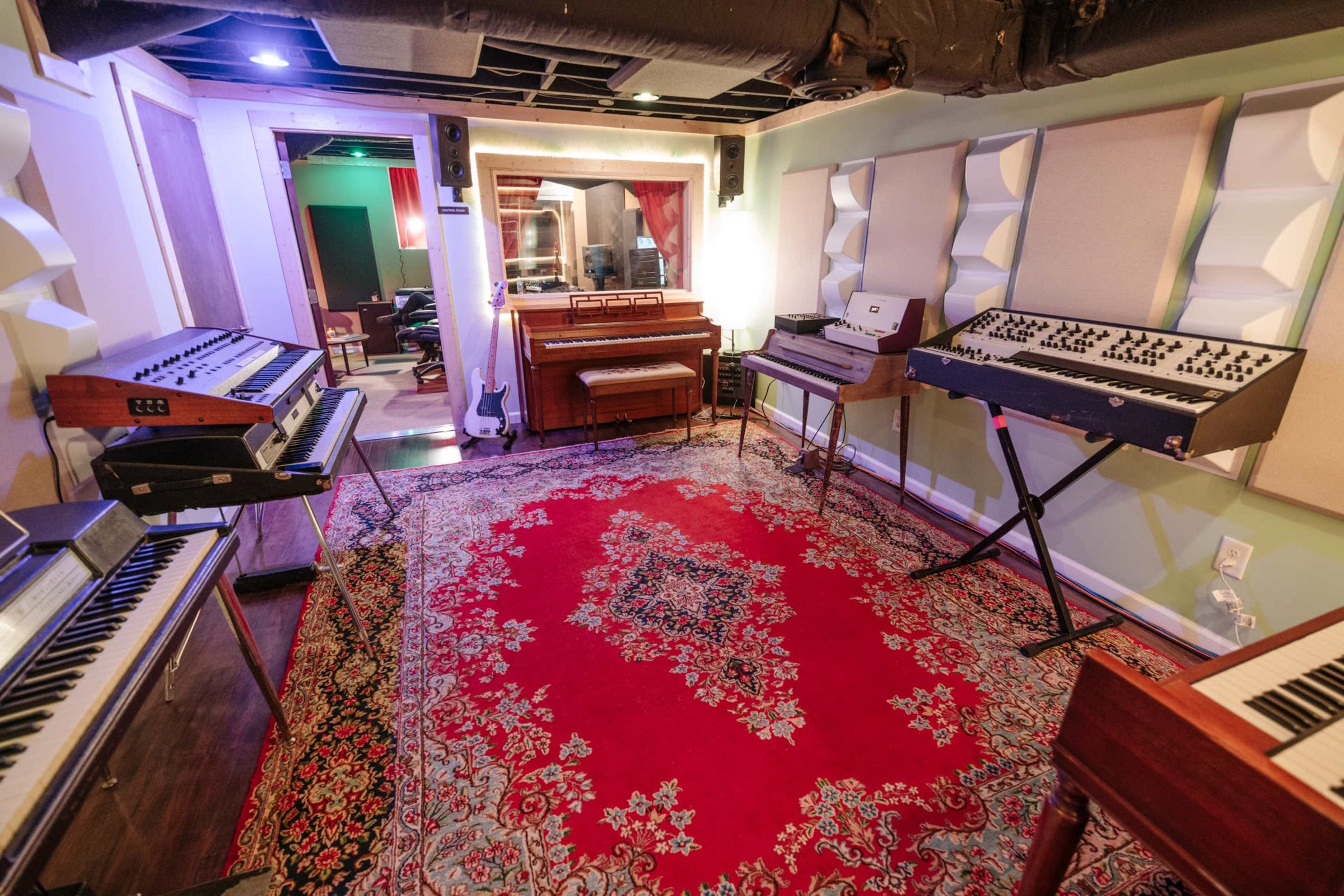 A music studio with various keyboards and synthesizers arranged around a red area rug, featuring a mirror and a recording area in the background.
