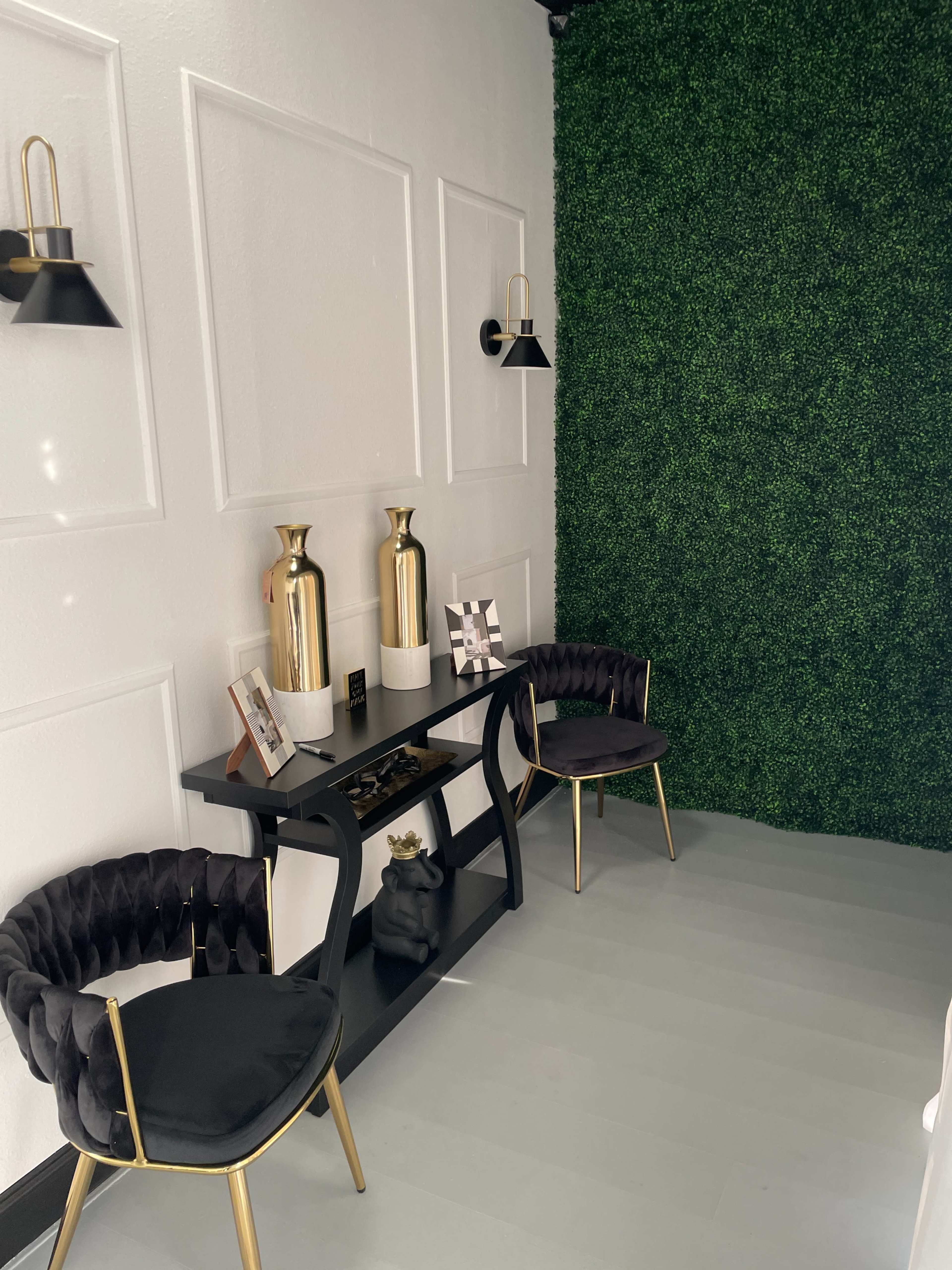 A modern interior space with a black and gold table, two upholstered chairs, and a green wall featuring artificial foliage.