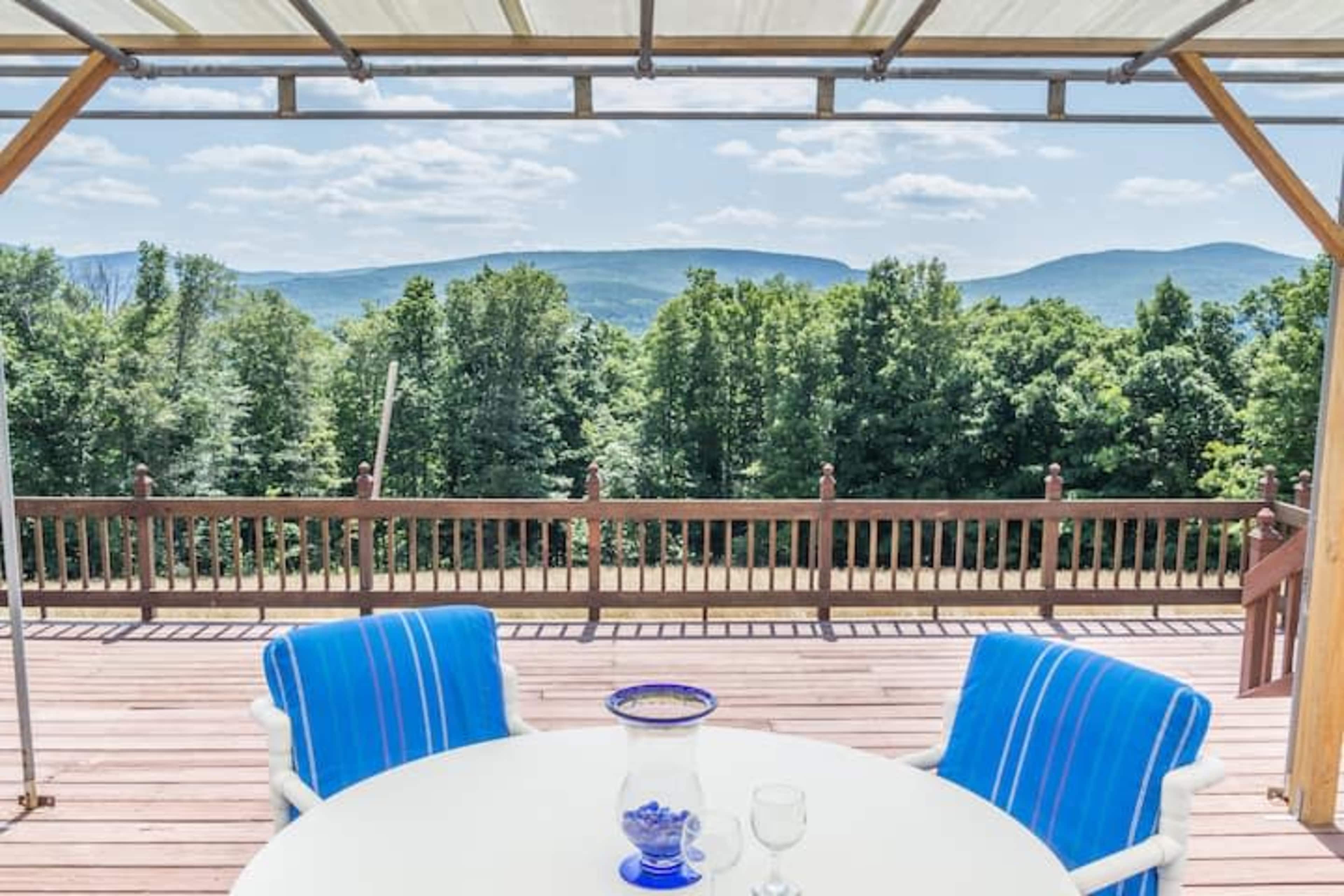 A wooden deck with a round table and two blue-striped chairs overlooks a green forest and distant mountains under a clear sky.