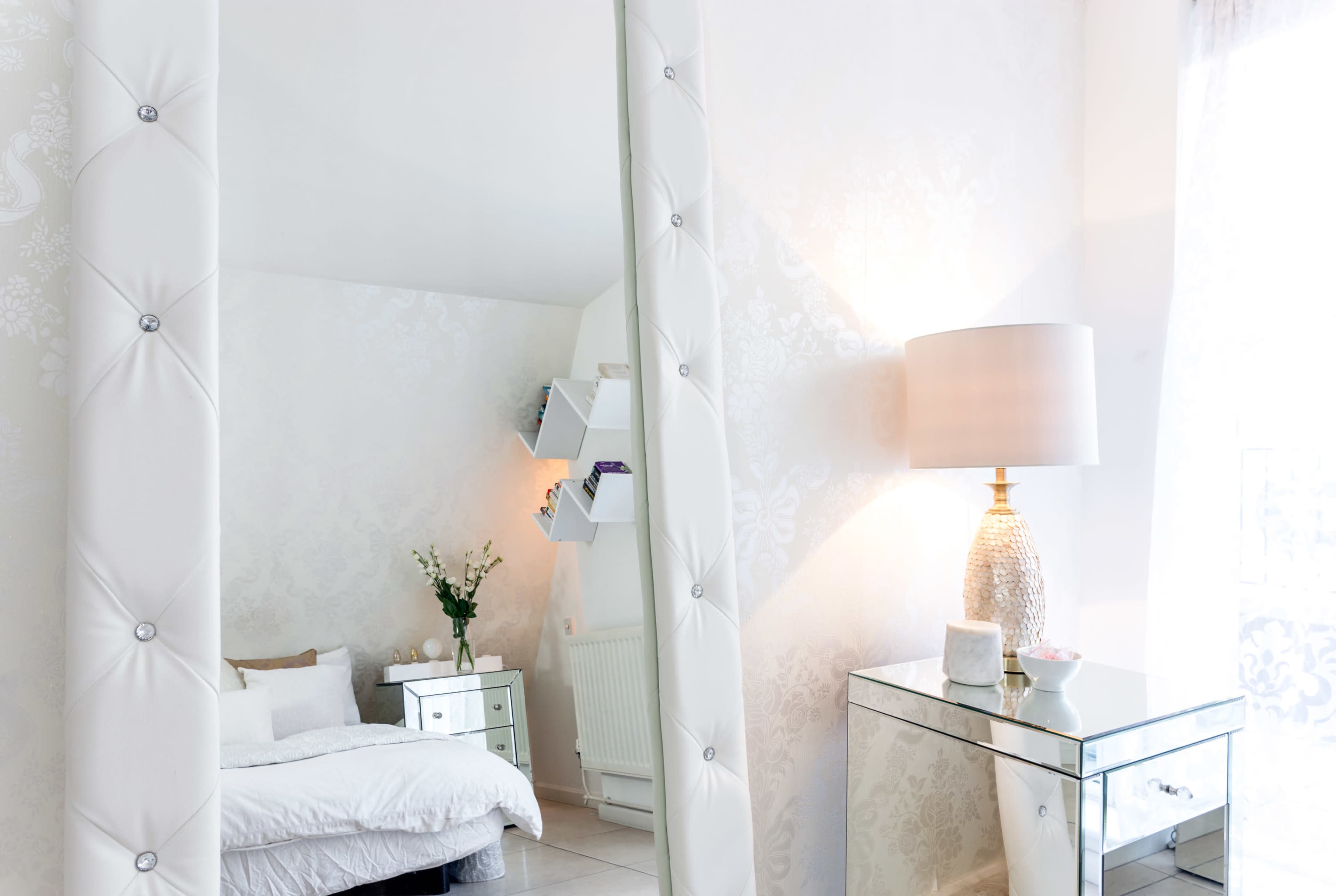 A bright bedroom features a mirrored nightstand, a large mirror, and a lamp beside a bed with white bedding.
