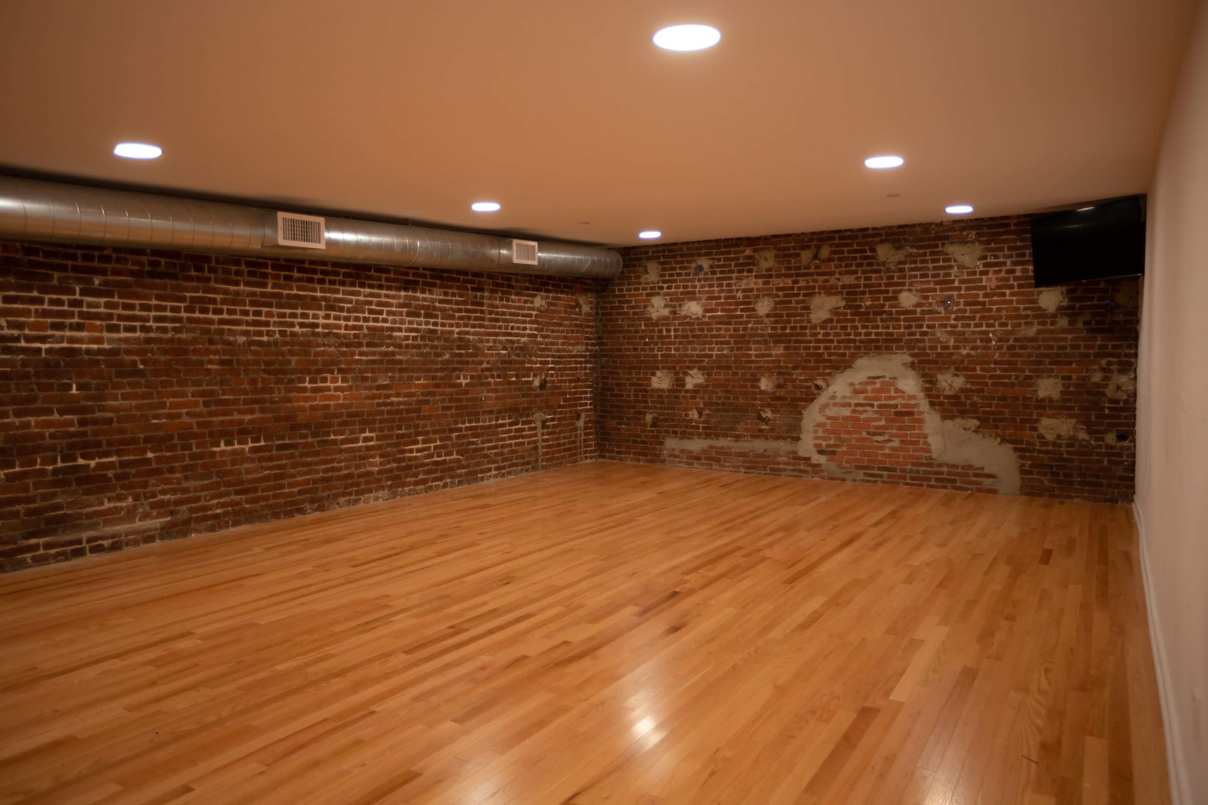 An empty room with hardwood flooring and exposed brick walls, featuring recessed lighting on the ceiling.