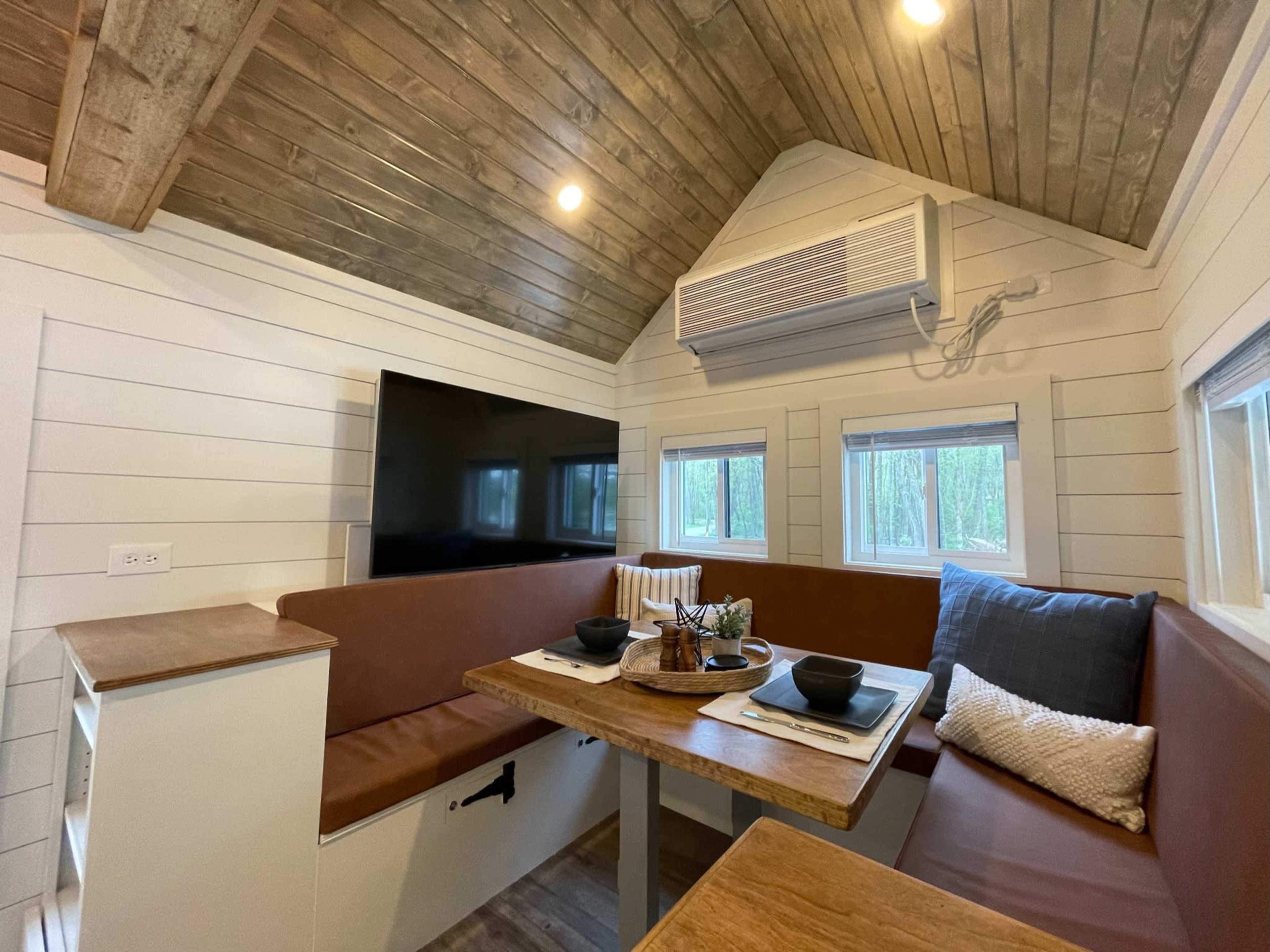 A cozy interior of a small cabin with a dining area featuring a wooden table and bench seating, complemented by a mounted TV and large windows.