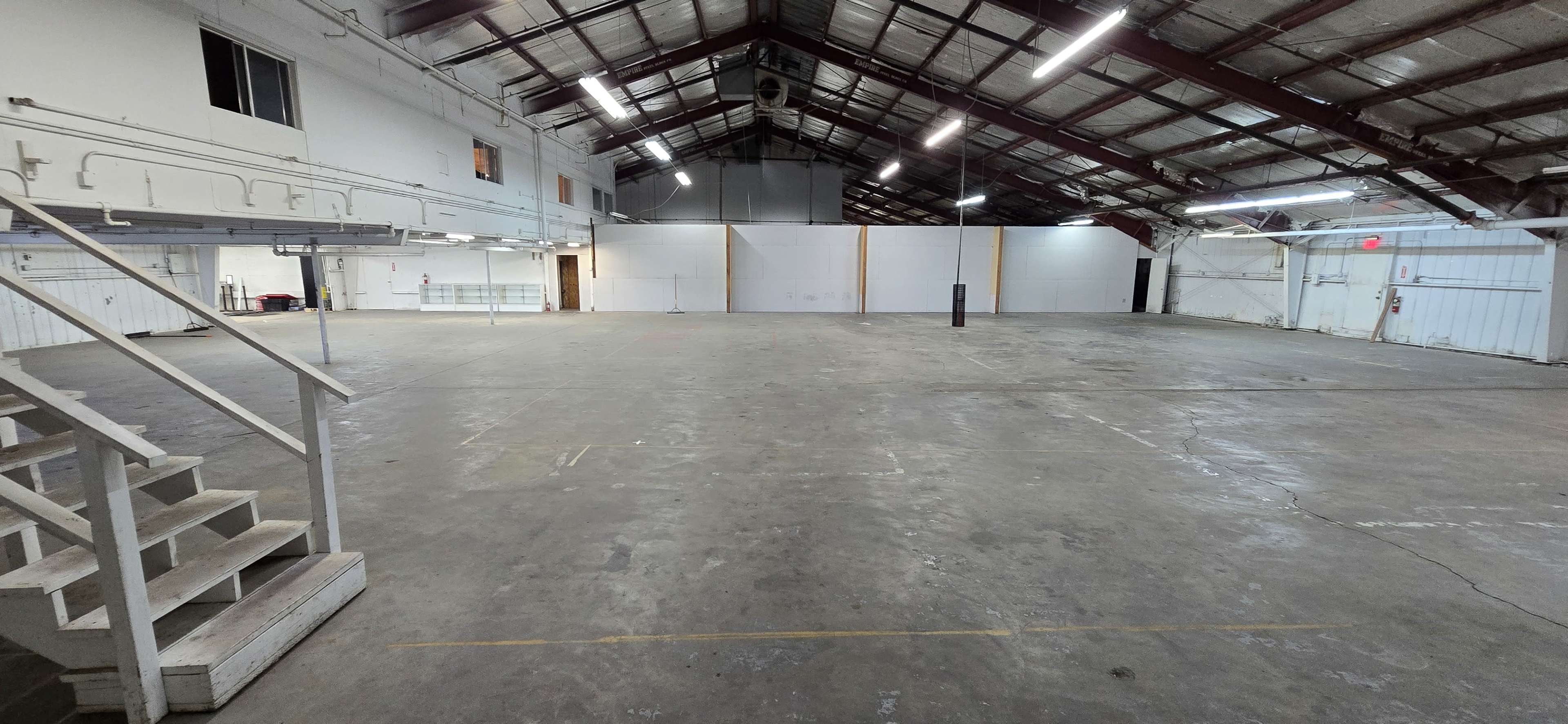 The image shows a spacious, empty industrial warehouse with high ceilings, exposed beams, and minimal furnishings.