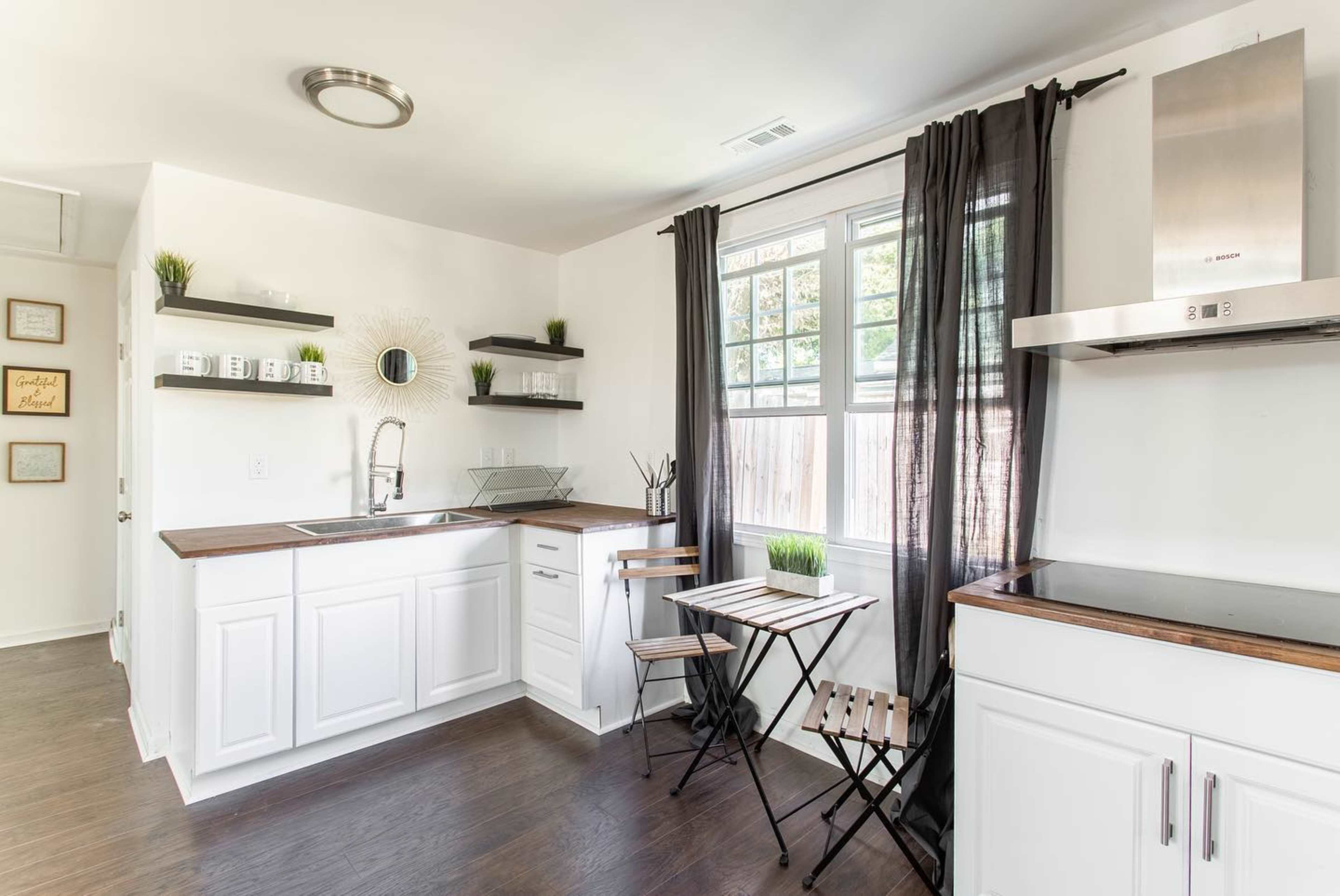 A modern kitchen with white cabinets, dark wooden countertops, shelves with plants and kitchenware, and a small dining table with chairs near a window.