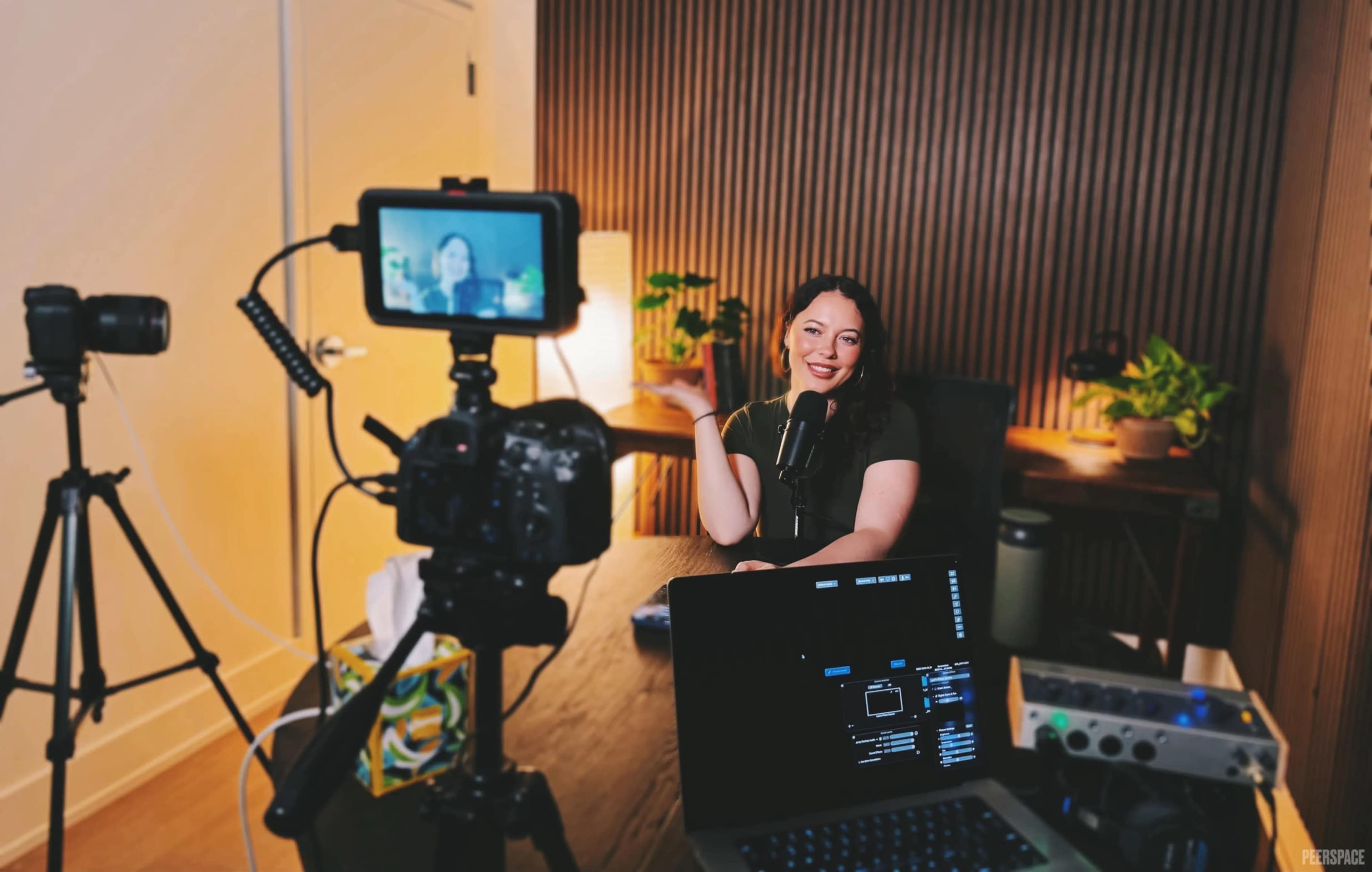 A woman is sitting at a desk with a microphone in front of her, while a camera and a laptop are set up to capture her on video.