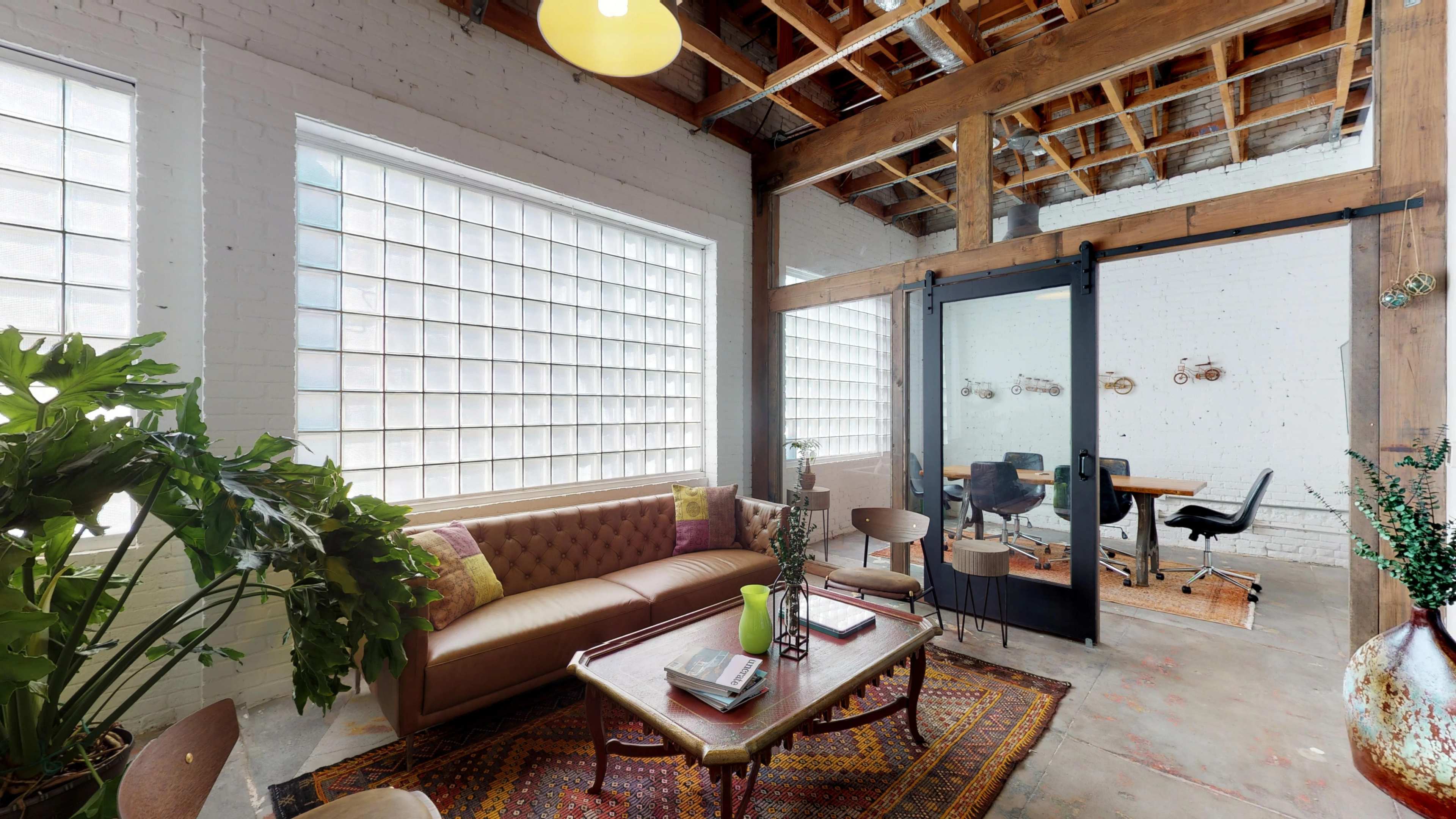 A stylish interior space featuring a brown leather sofa, a wooden coffee table, and a glass-brick window, with a separate meeting area visible through a glass door.