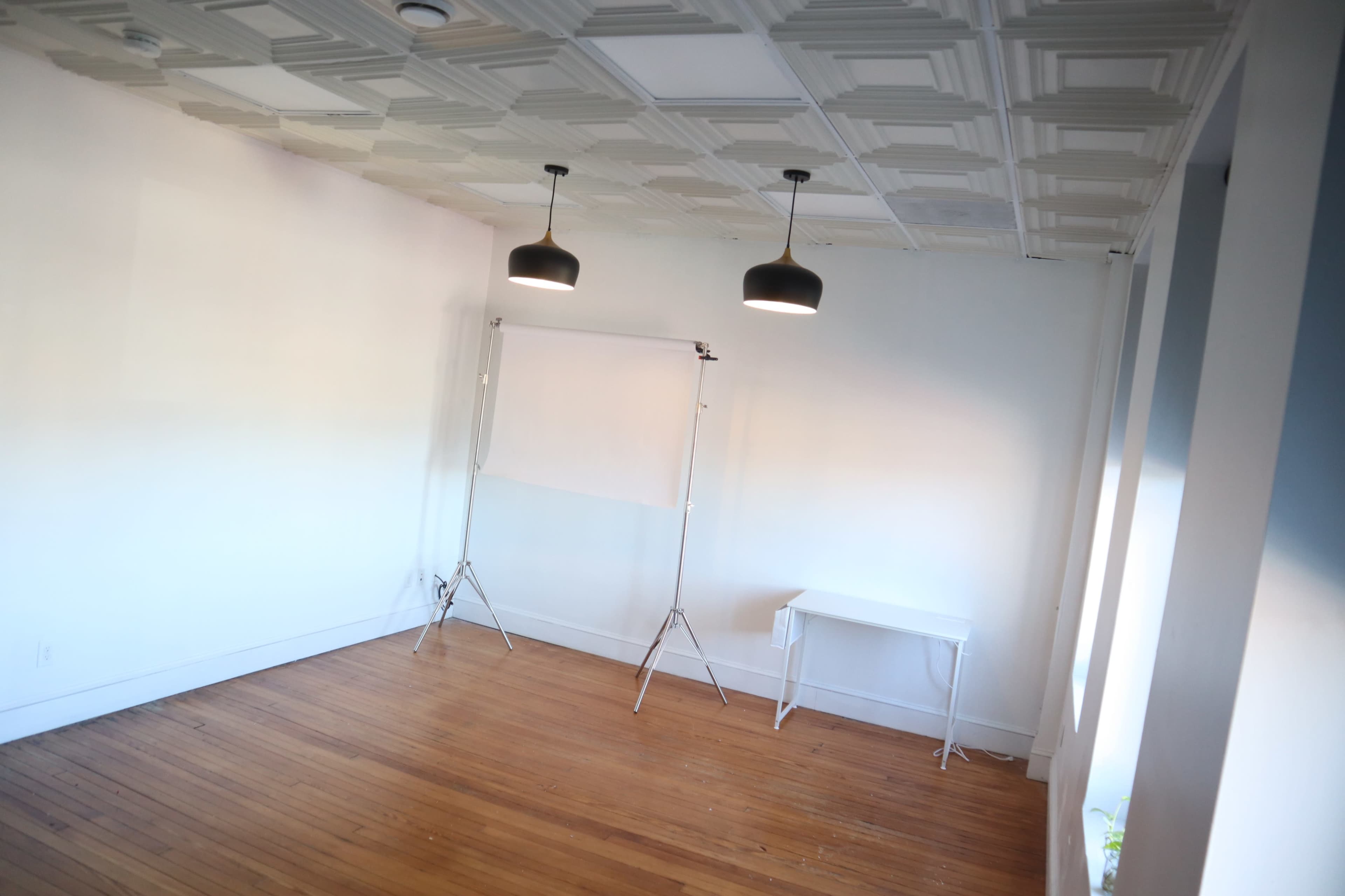 A spacious, empty room with wooden flooring, two pendant lights hanging from the ceiling, and a white backdrop stand near a small white table.