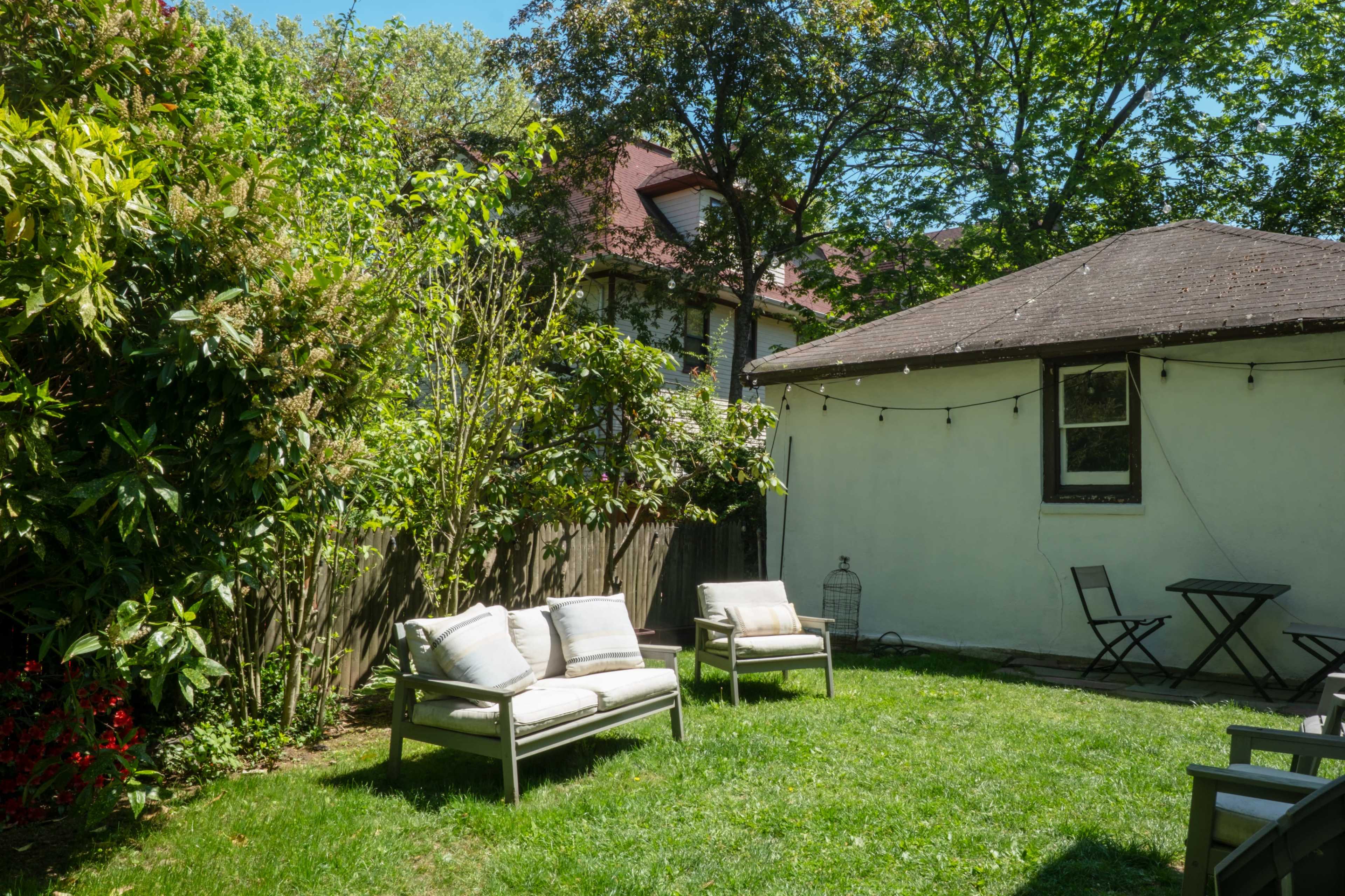 The image shows a green backyard with two chairs and a small table, surrounded by trees and a white wall building.