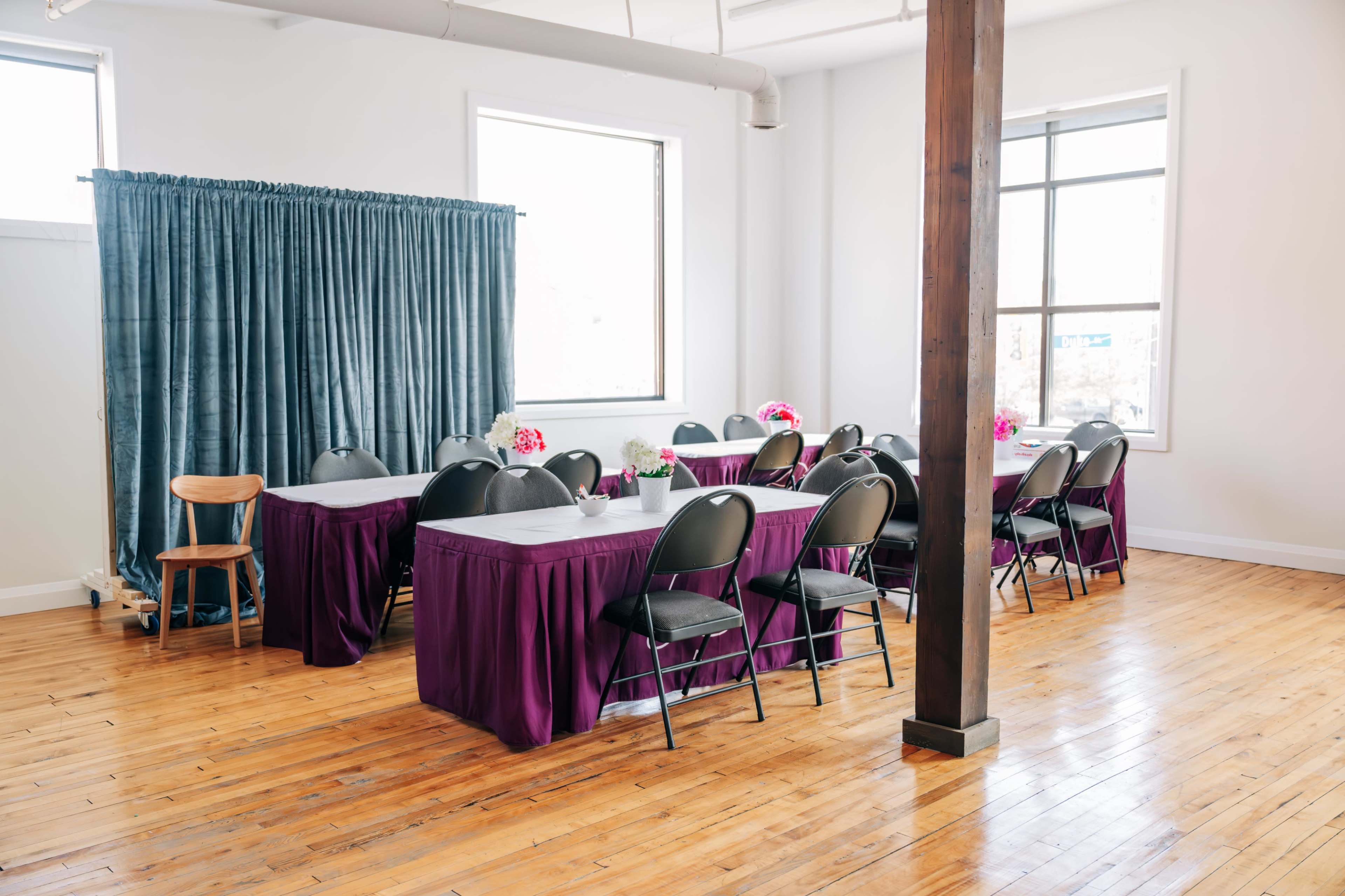 A well-lit room features several tables covered in purple tablecloths, arranged with black chairs and floral centerpieces.