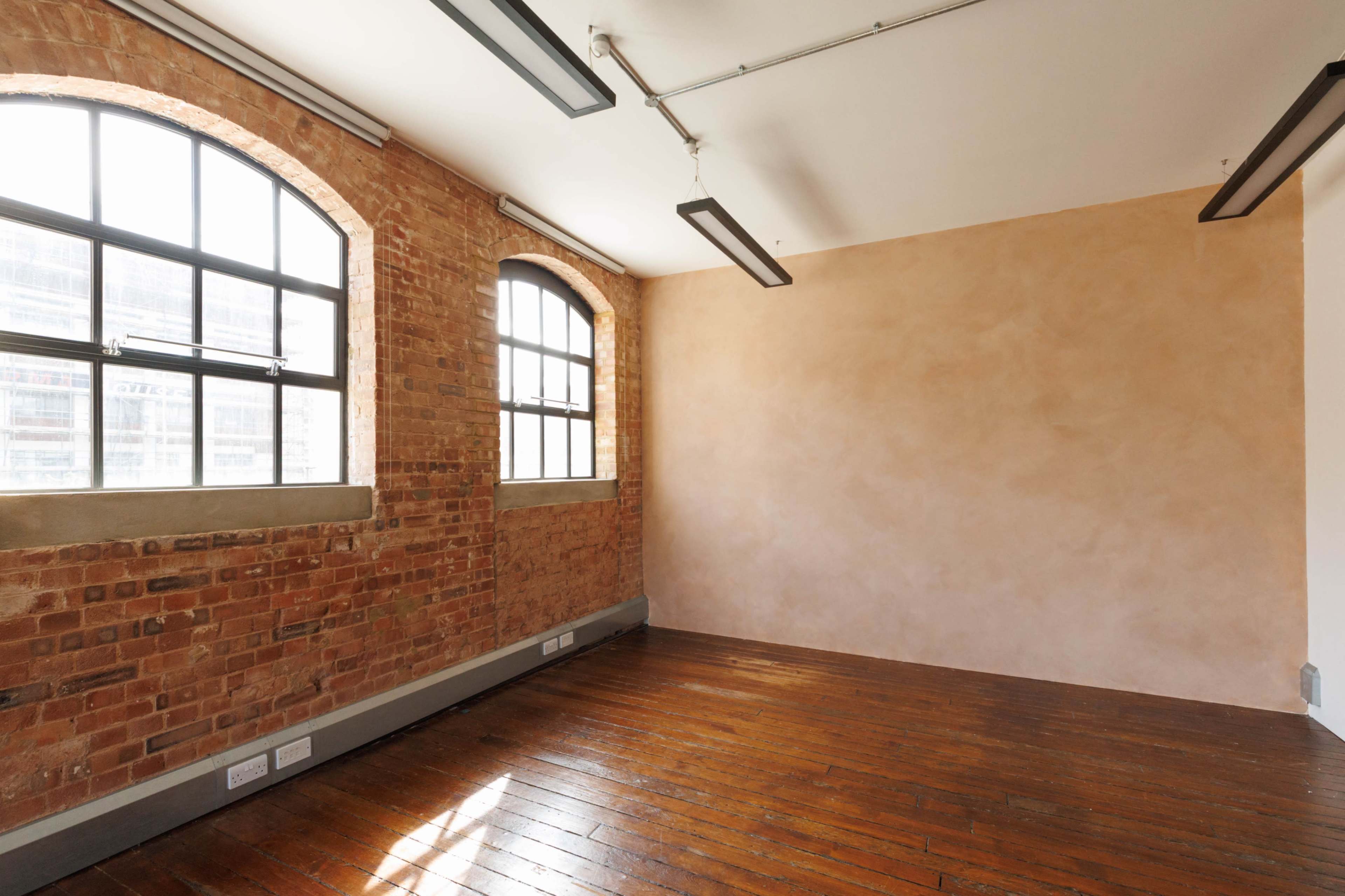 The image shows a spacious, empty room with exposed brick walls, large windows, and wooden flooring.