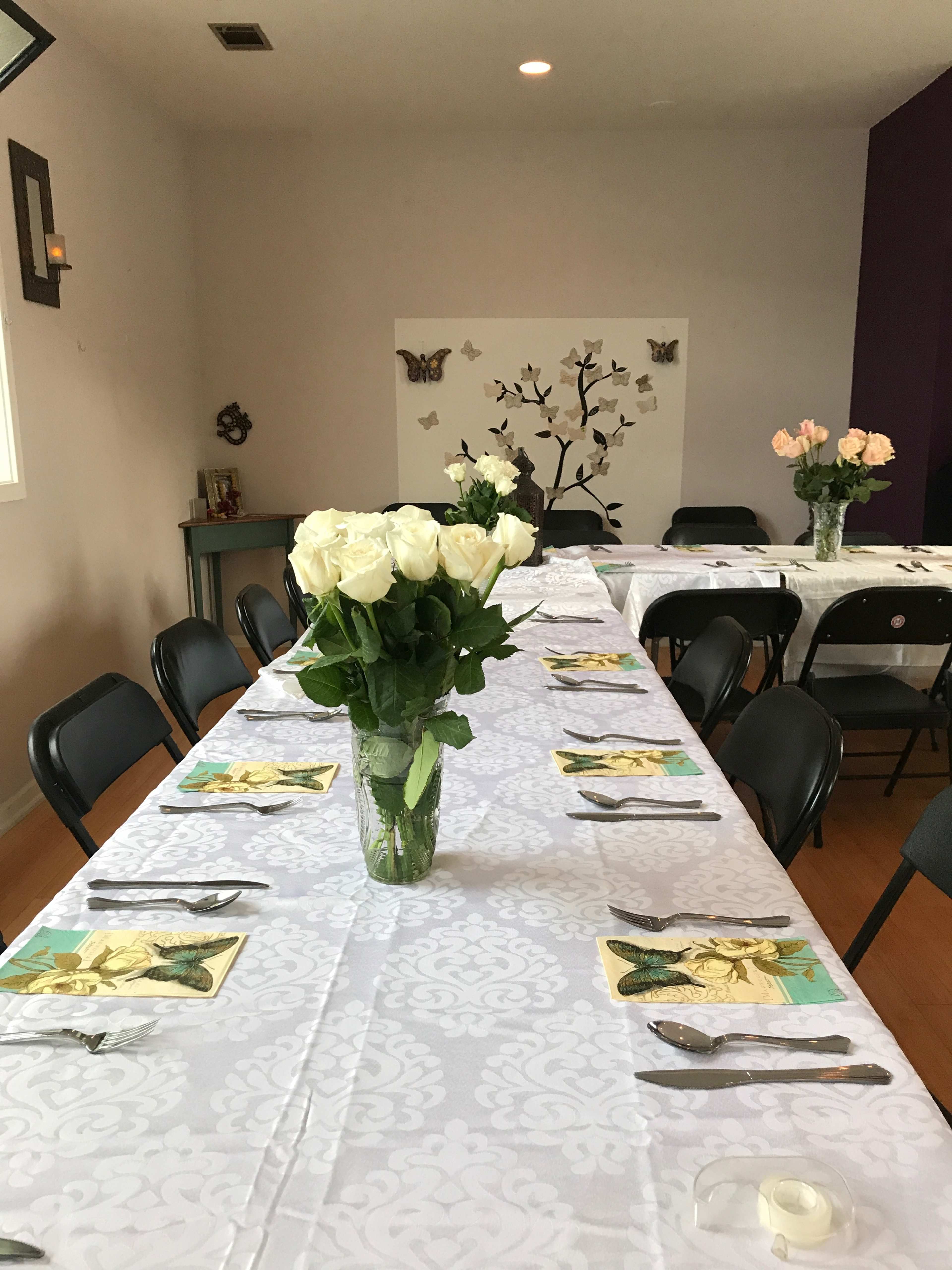 A long, neatly set dining table with white tablecloths, silverware, and a vase of white roses sits in a room decorated with a butterfly-themed wall art.