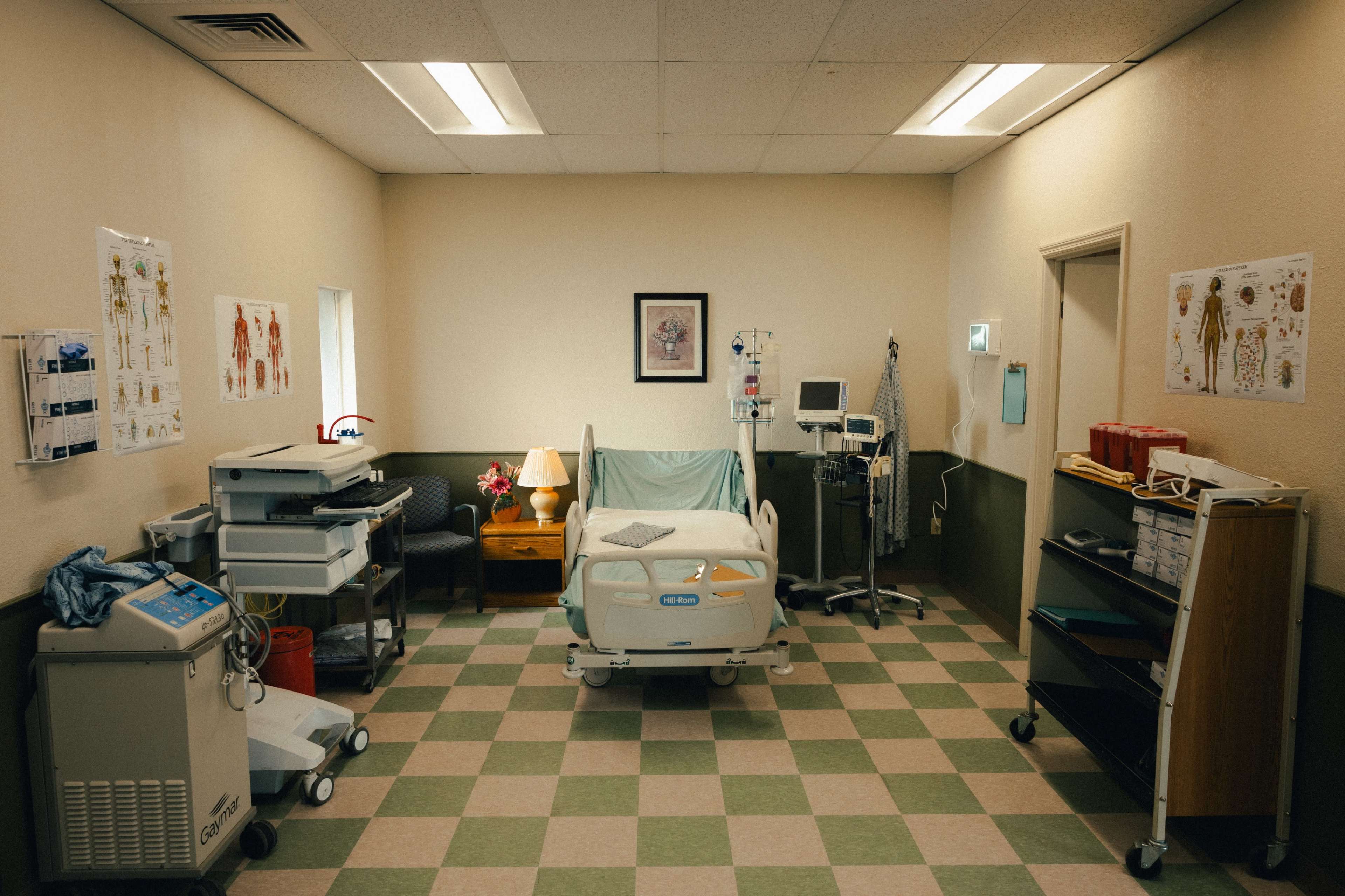 A sterile hospital room with a bed, medical equipment, a lamp on a small table, and charts on the walls.