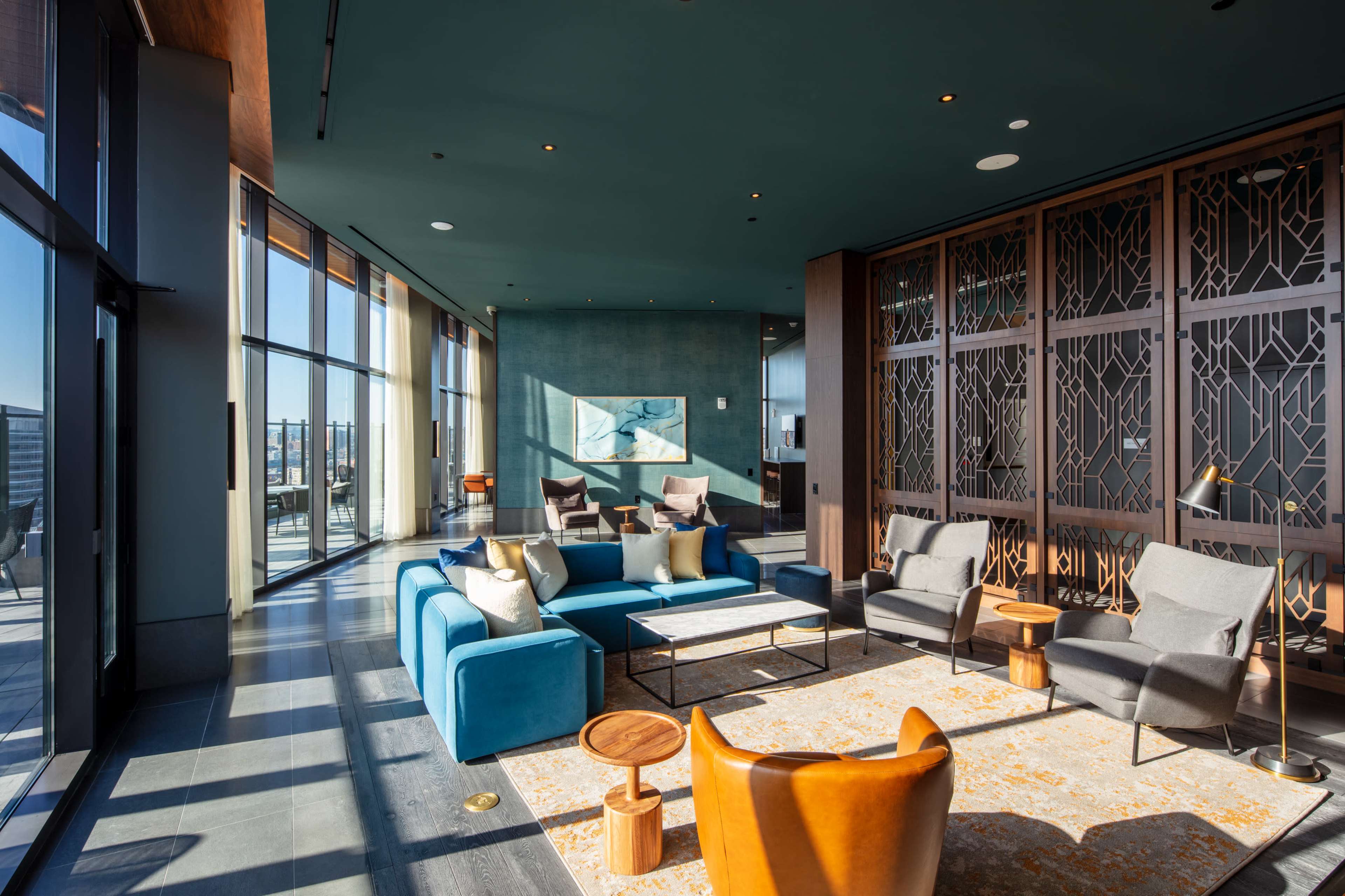 The image shows a modern lounge area with large windows, featuring a blue sectional sofa, gray armchairs, small wooden tables, and decorative artwork on the walls.