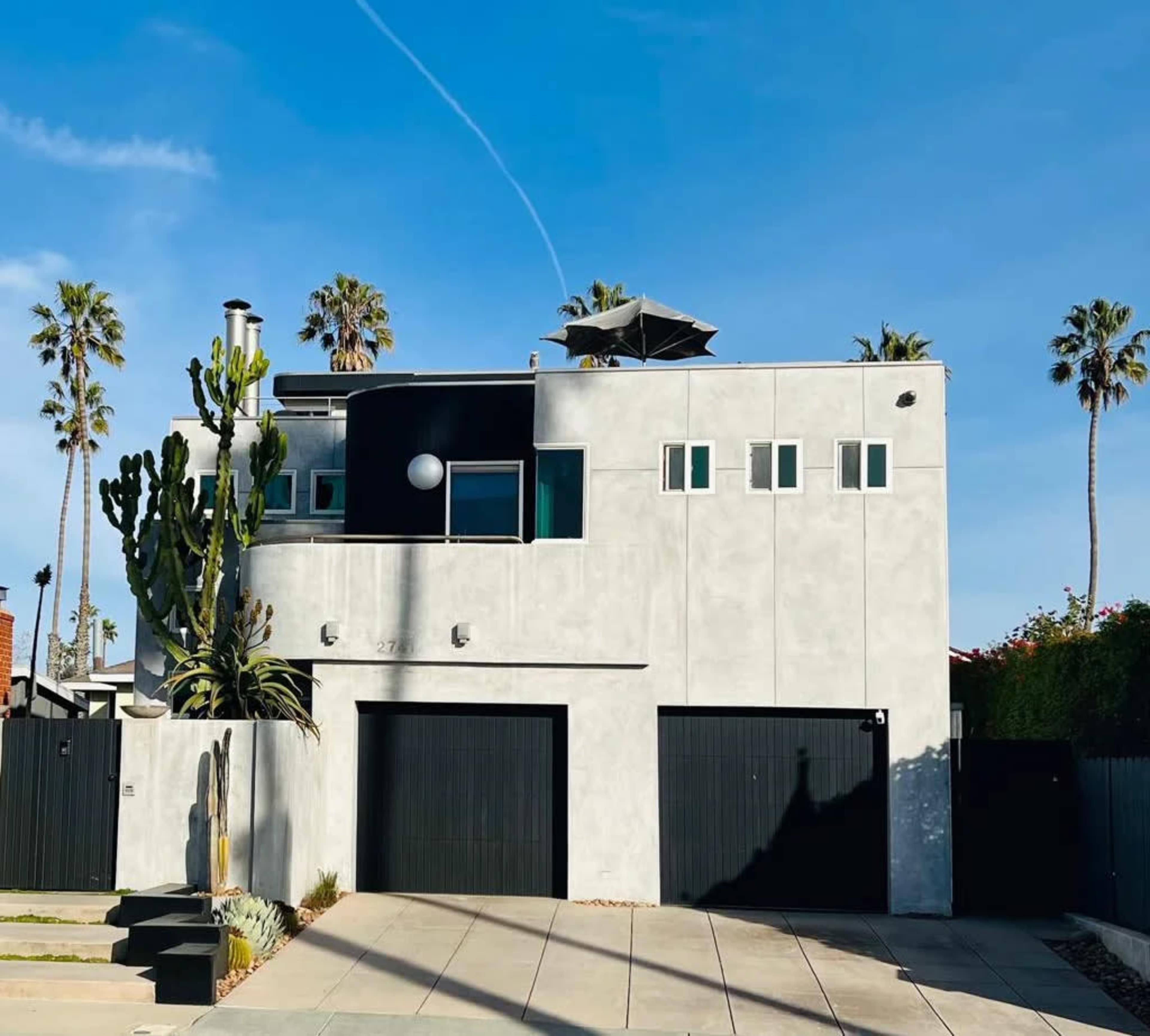 A modern two-story house with a concrete exterior, featuring large windows, a flat roof, and two black garage doors, surrounded by palm trees.