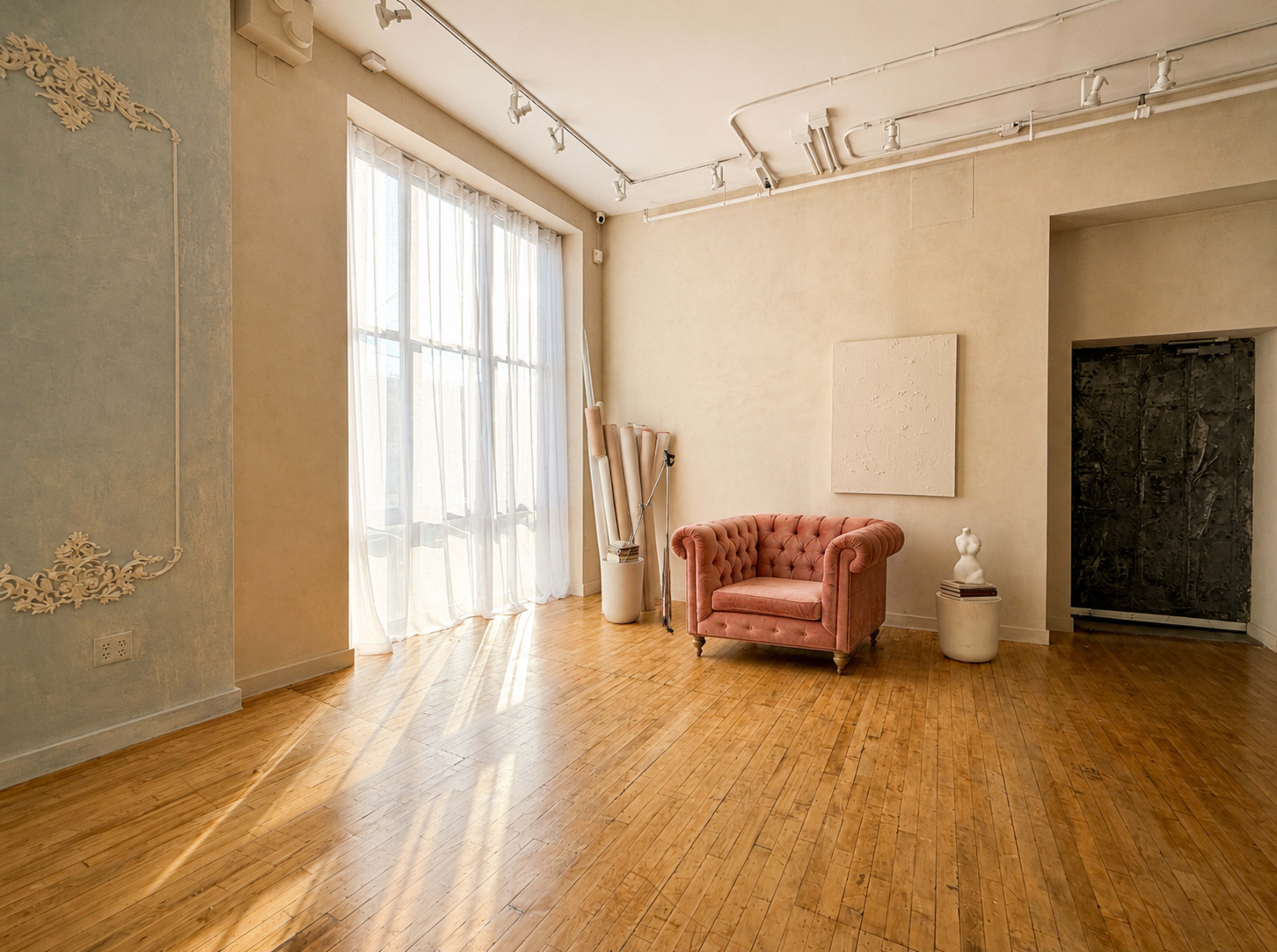 The image shows a sunlit interior room with a pink tufted armchair, wooden flooring, and large windows draped with sheer curtains.