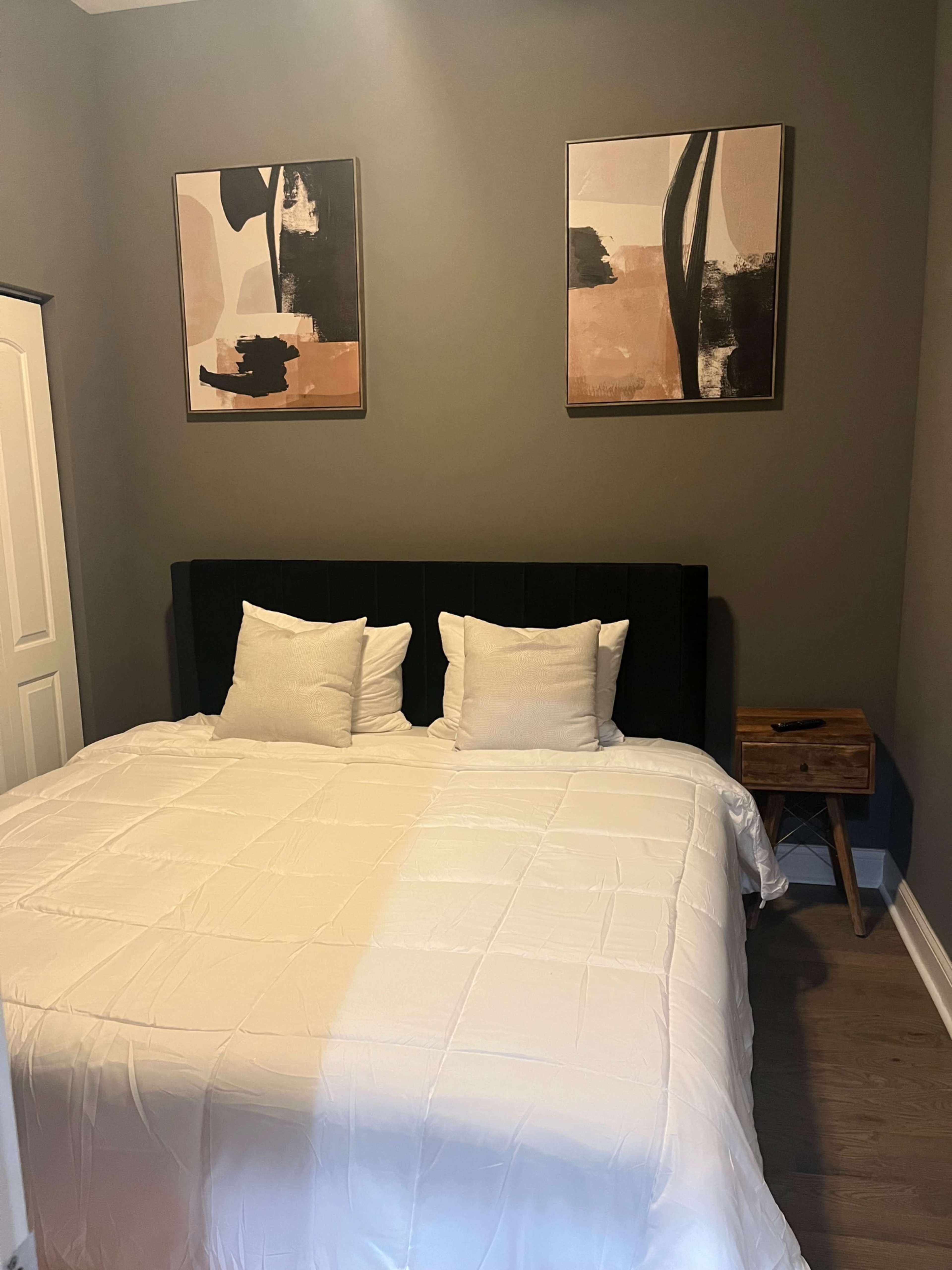 A neatly made bed with white bedding, two pillows, and abstract art pieces hanging on the gray wall above it, alongside a small wooden nightstand.