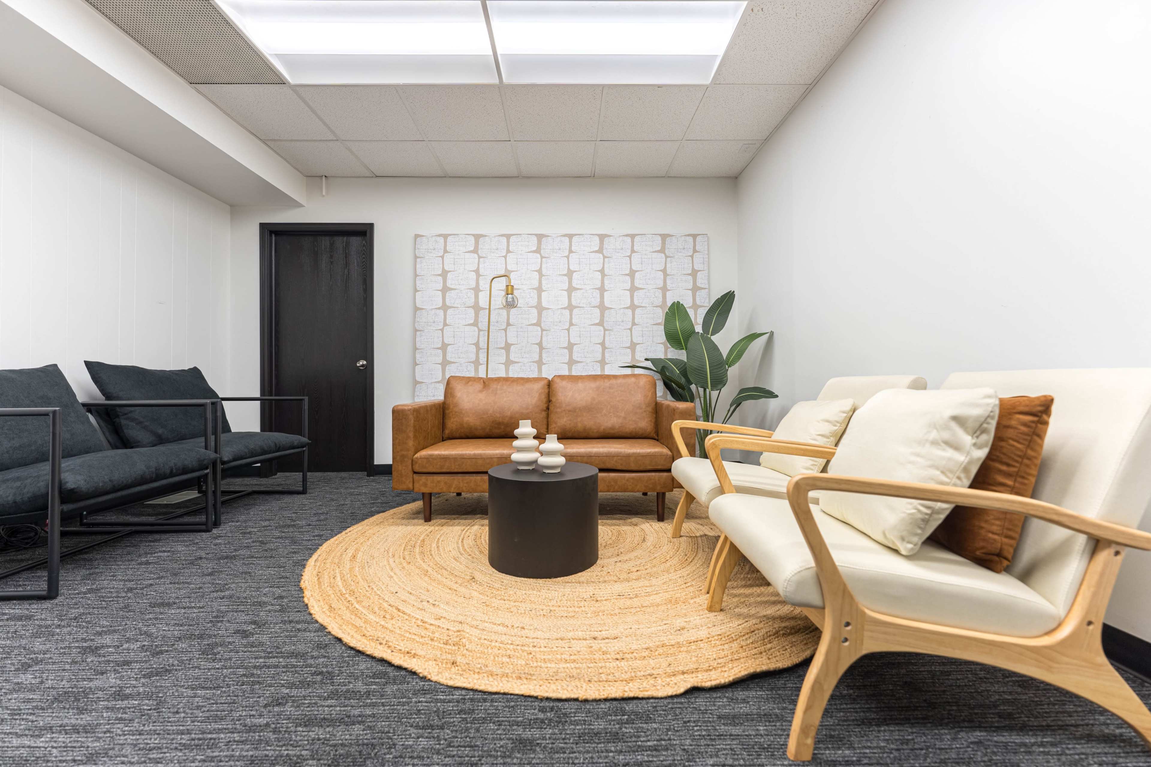 The image shows a modern waiting area with a tan leather sofa, light-colored armchairs, a round black table, and a large circular rug.