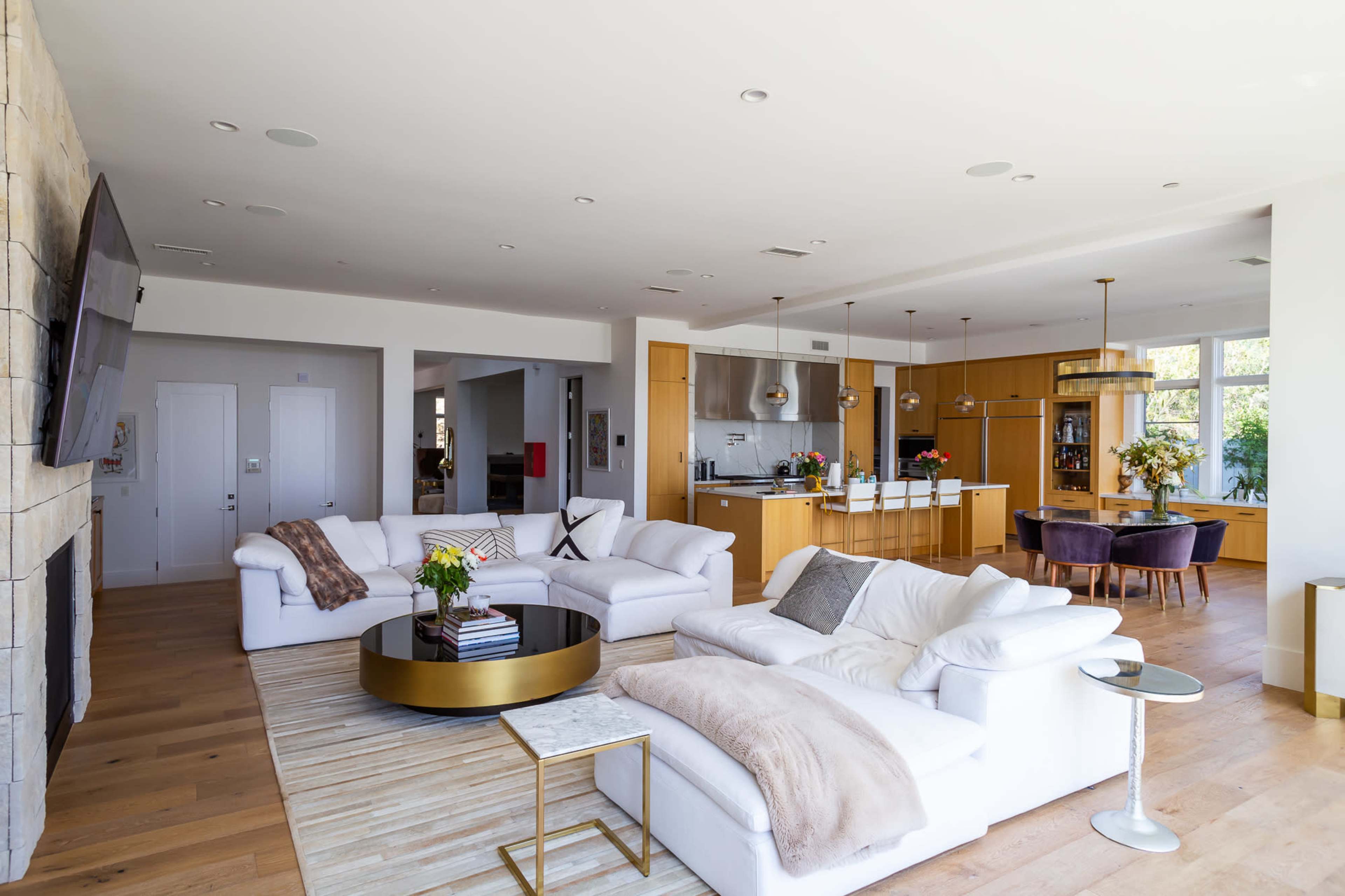 The image shows a modern living area with white sofas, a round coffee table, and an open kitchen, featuring wooden floors and large windows.