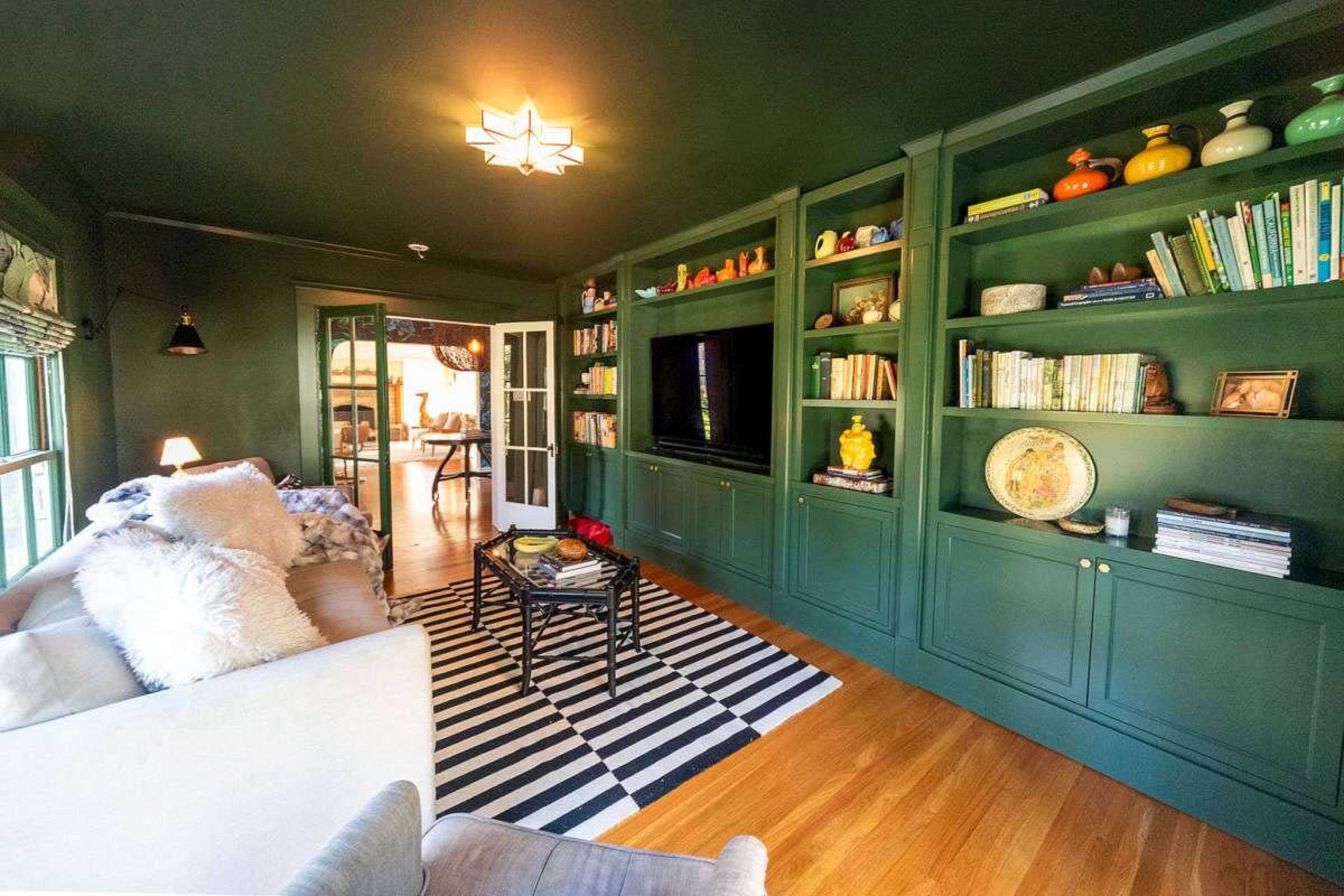 The image shows a cozy living room with dark green walls, built-in shelving filled with books and decorative items, a striped area rug, and a couch facing a television.