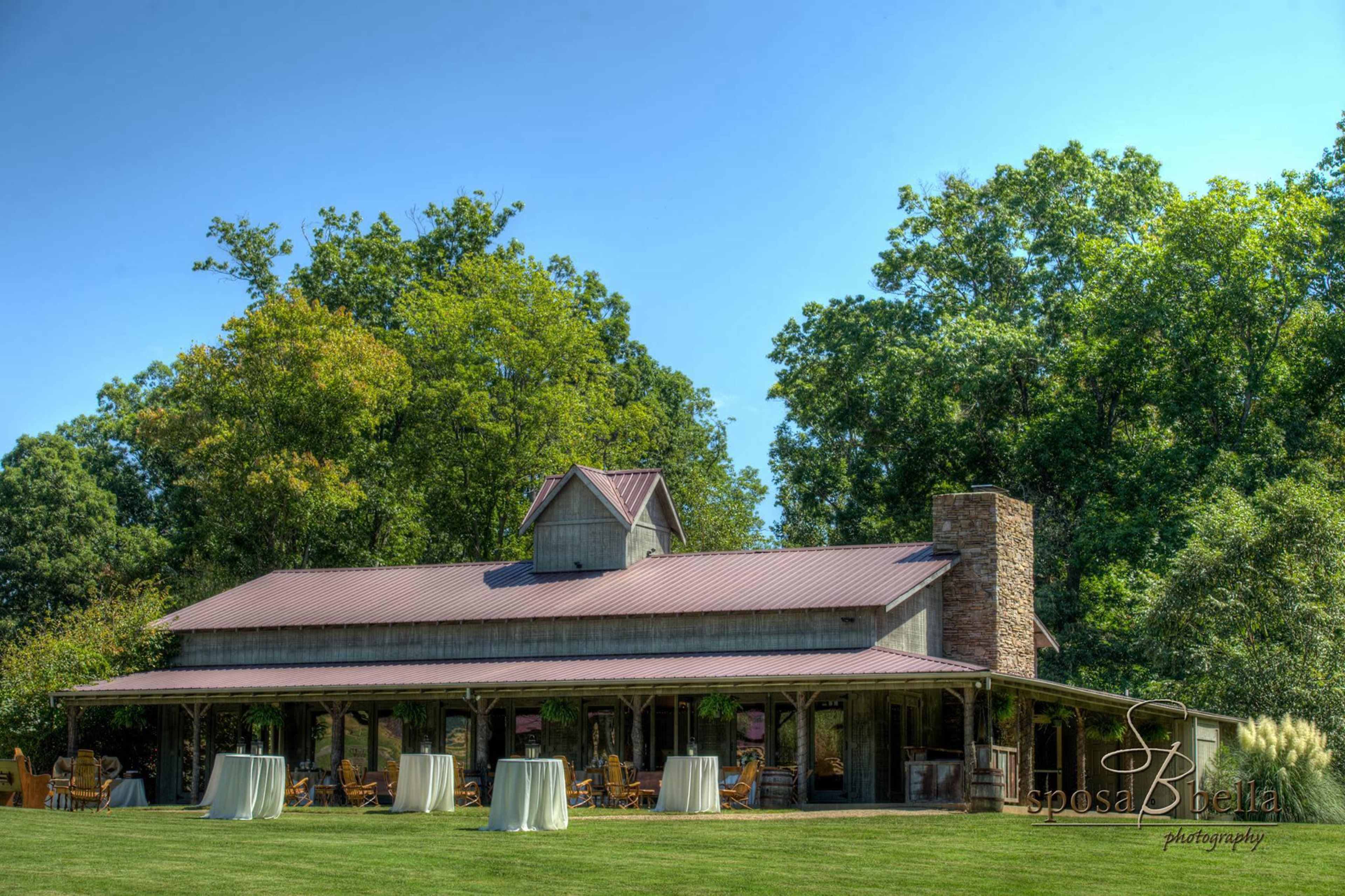A rustic venue features a wooden structure with a metal roof, surrounded by greenery and set with tables on the lawn.