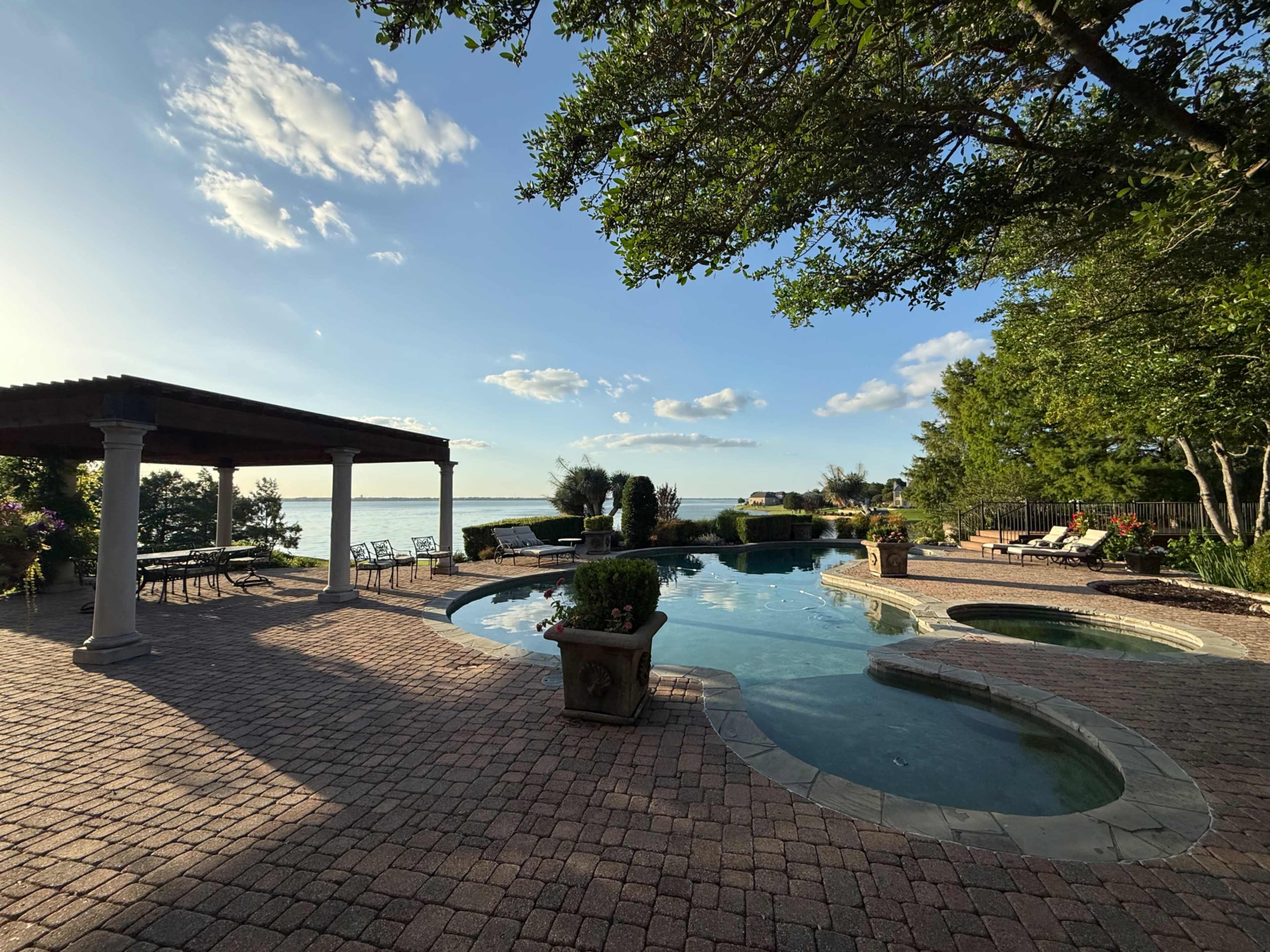 The image shows a backyard with a swimming pool, spa area, and seating surrounded by trees and a view of a lake under a partly cloudy sky.