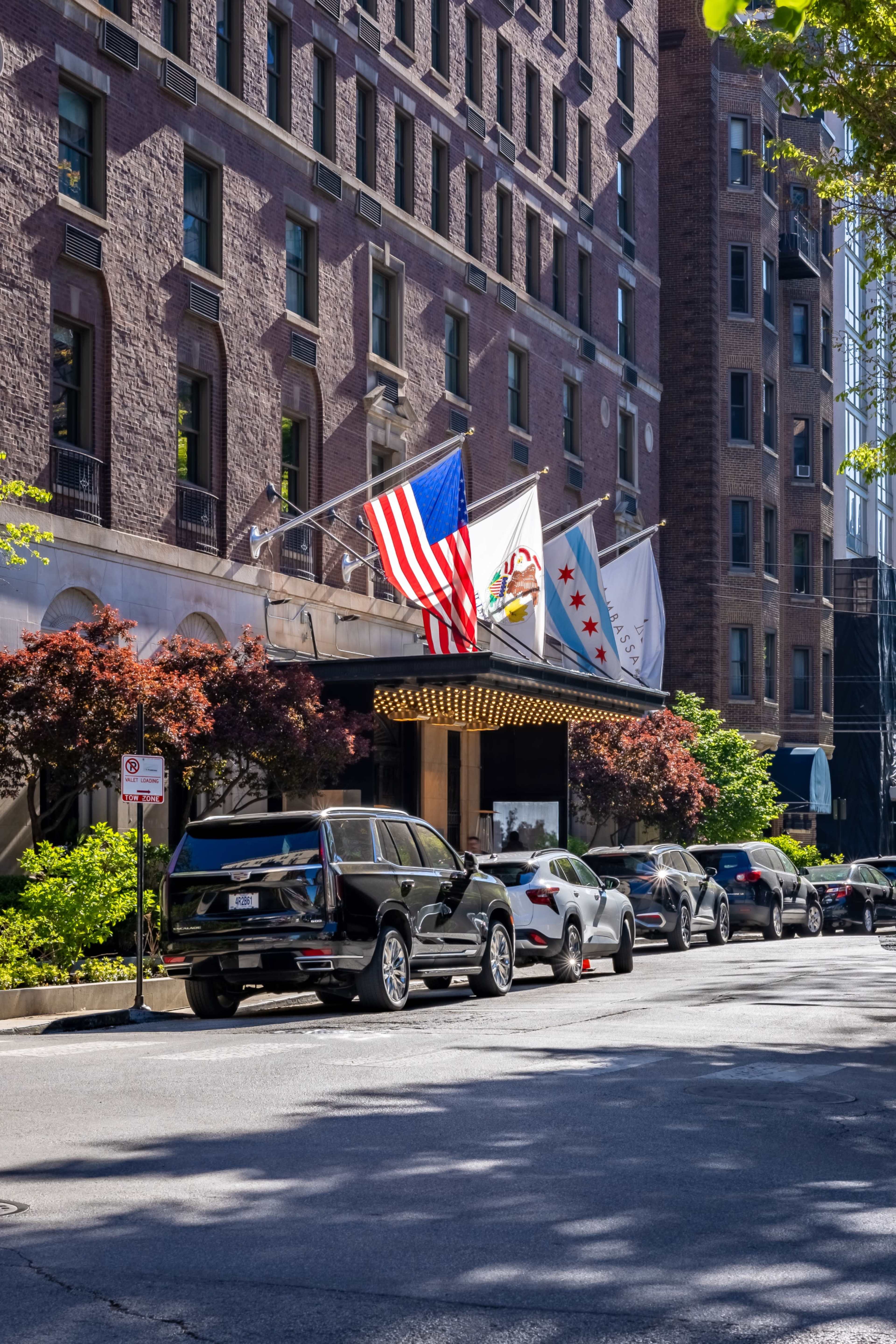 The image shows a brick hotel facade with an entrance canopy, flanked by several flags, and lined with parked cars along the street.