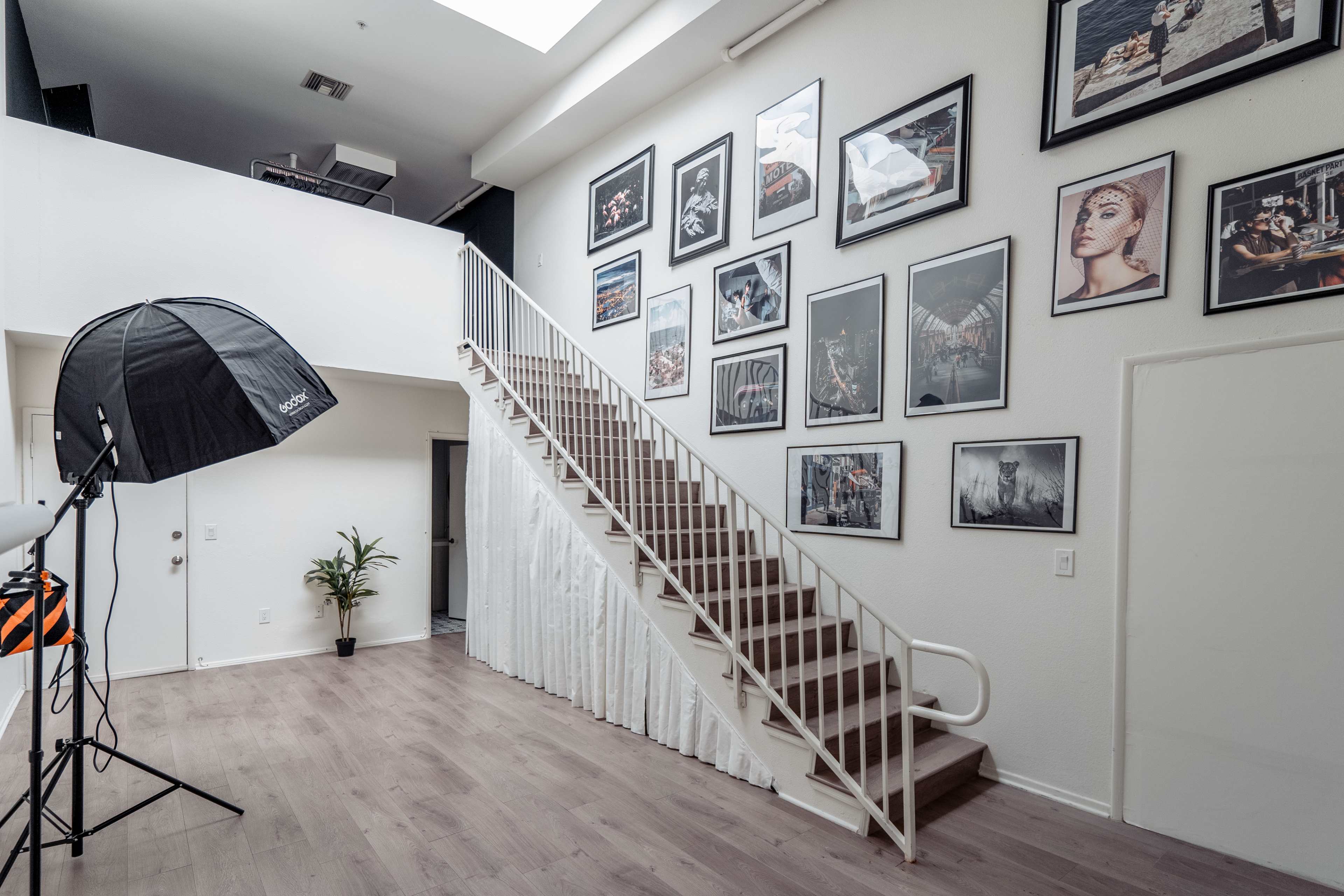 A bright interior features a staircase leading to an upper level, adorned with framed photographs on the walls, with a photography lighting setup in the foreground.