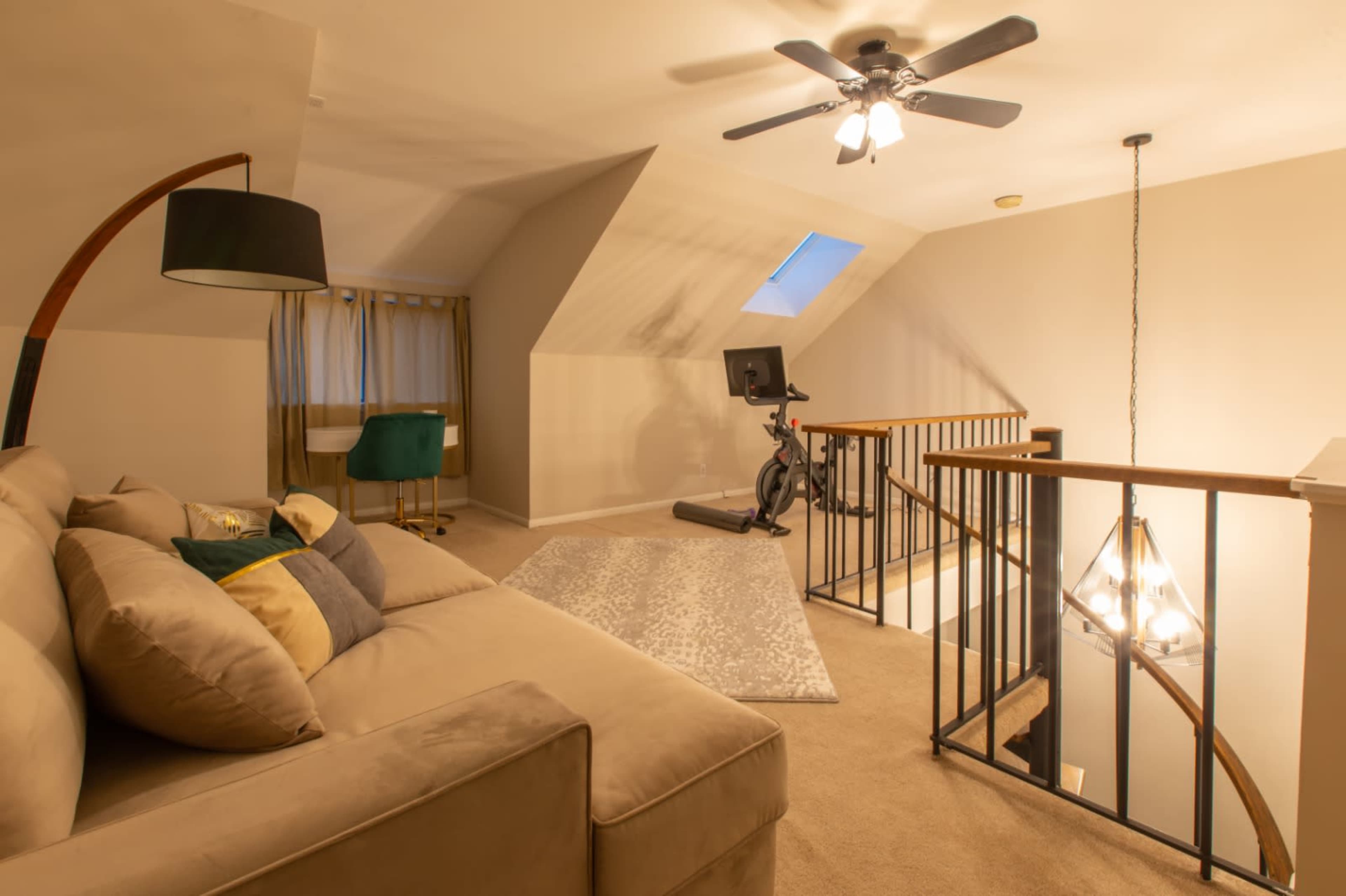 A cozy loft area featuring a sectional sofa, a standing lamp, a small desk with a green chair, a stationary bike, and a chandelier above the staircase.