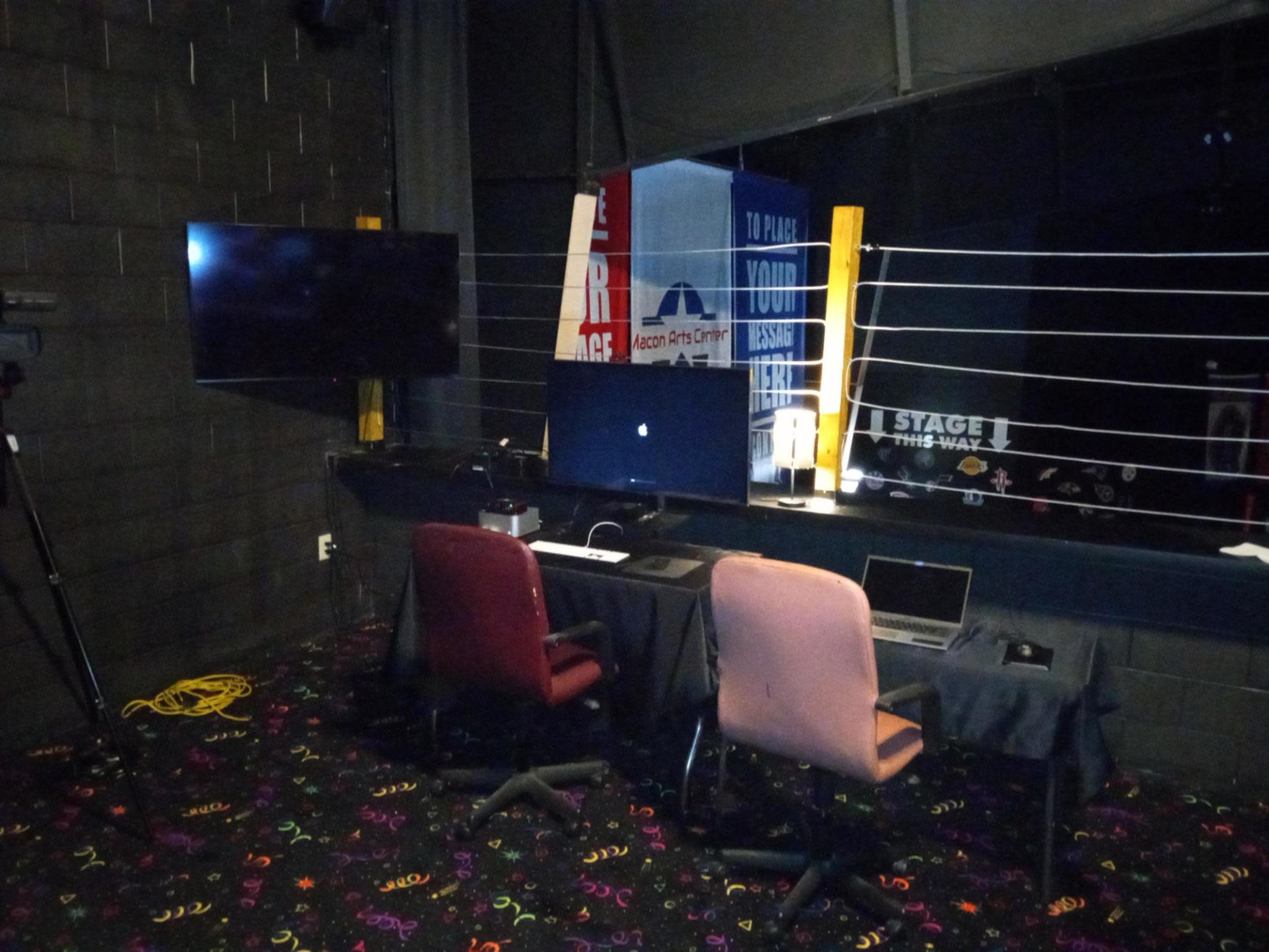 The image shows a dimly lit room with two desks equipped with laptops and monitors, along with a camera setup in the corner.
