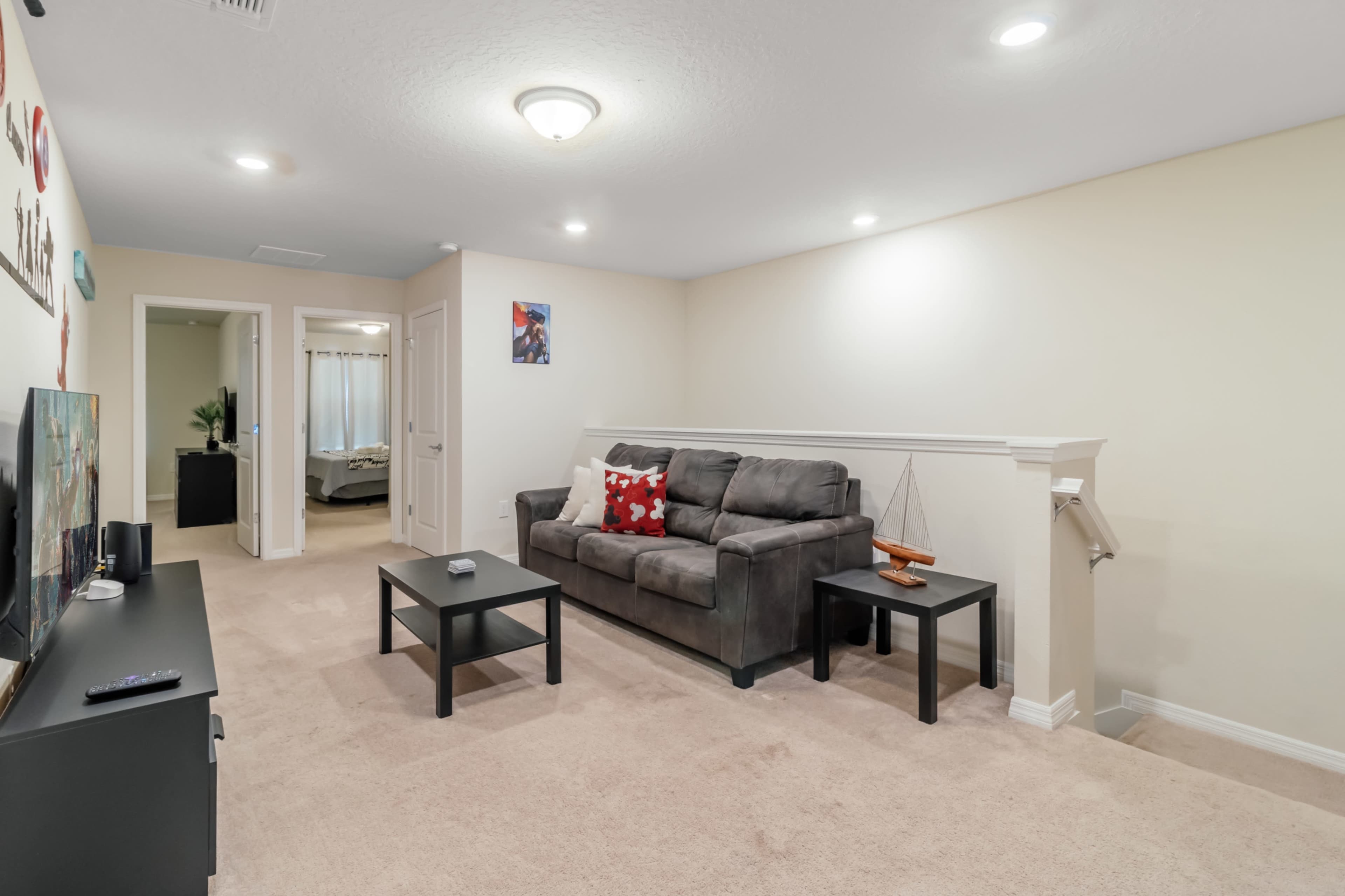 A living room features a gray sofa with red decorative pillows and two black coffee tables, with a television on a stand and a doorway leading to a bedroom area.