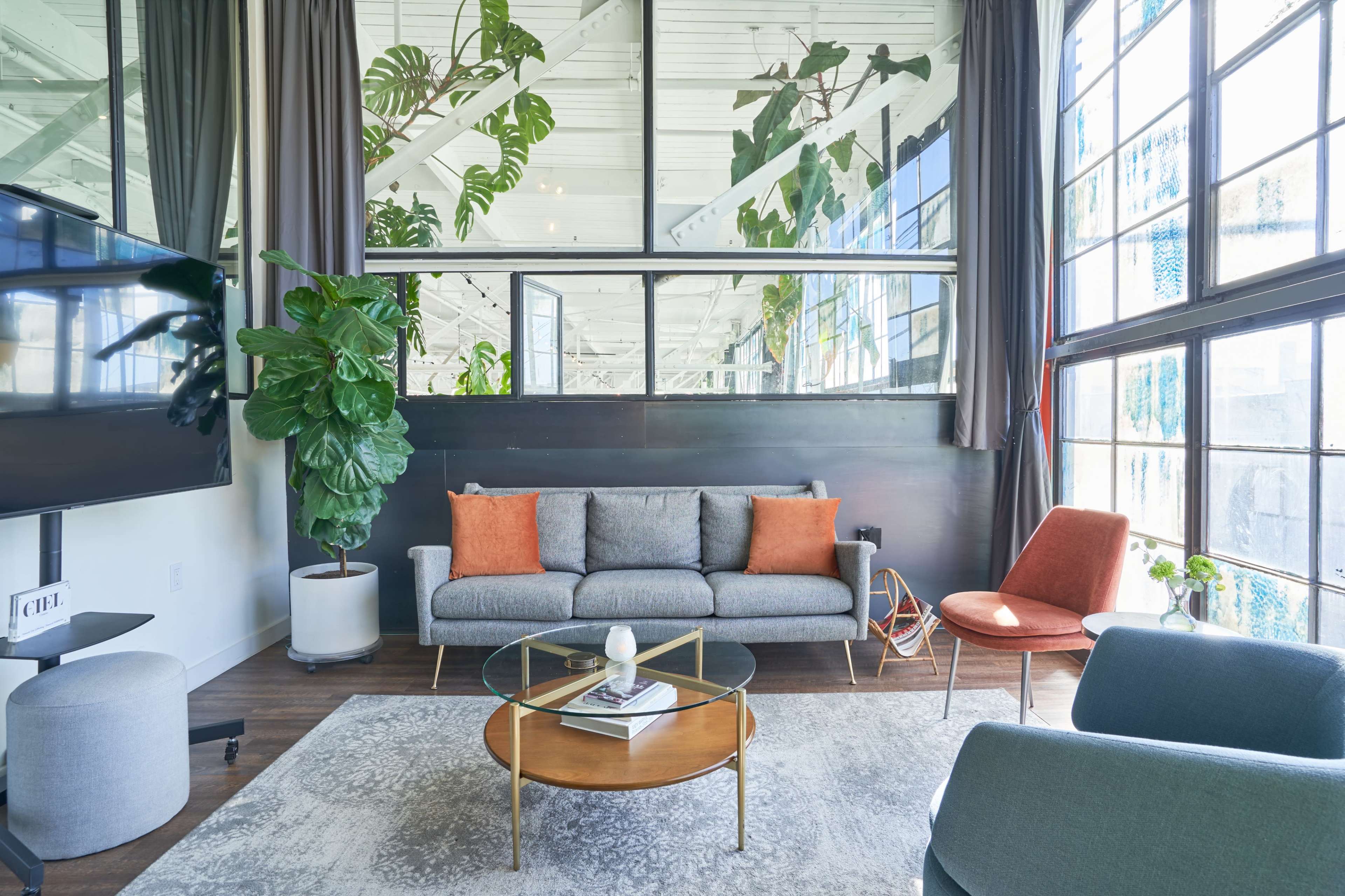 A modern living room with large windows, a gray sofa with orange accent pillows, a round coffee table, and indoor plants.