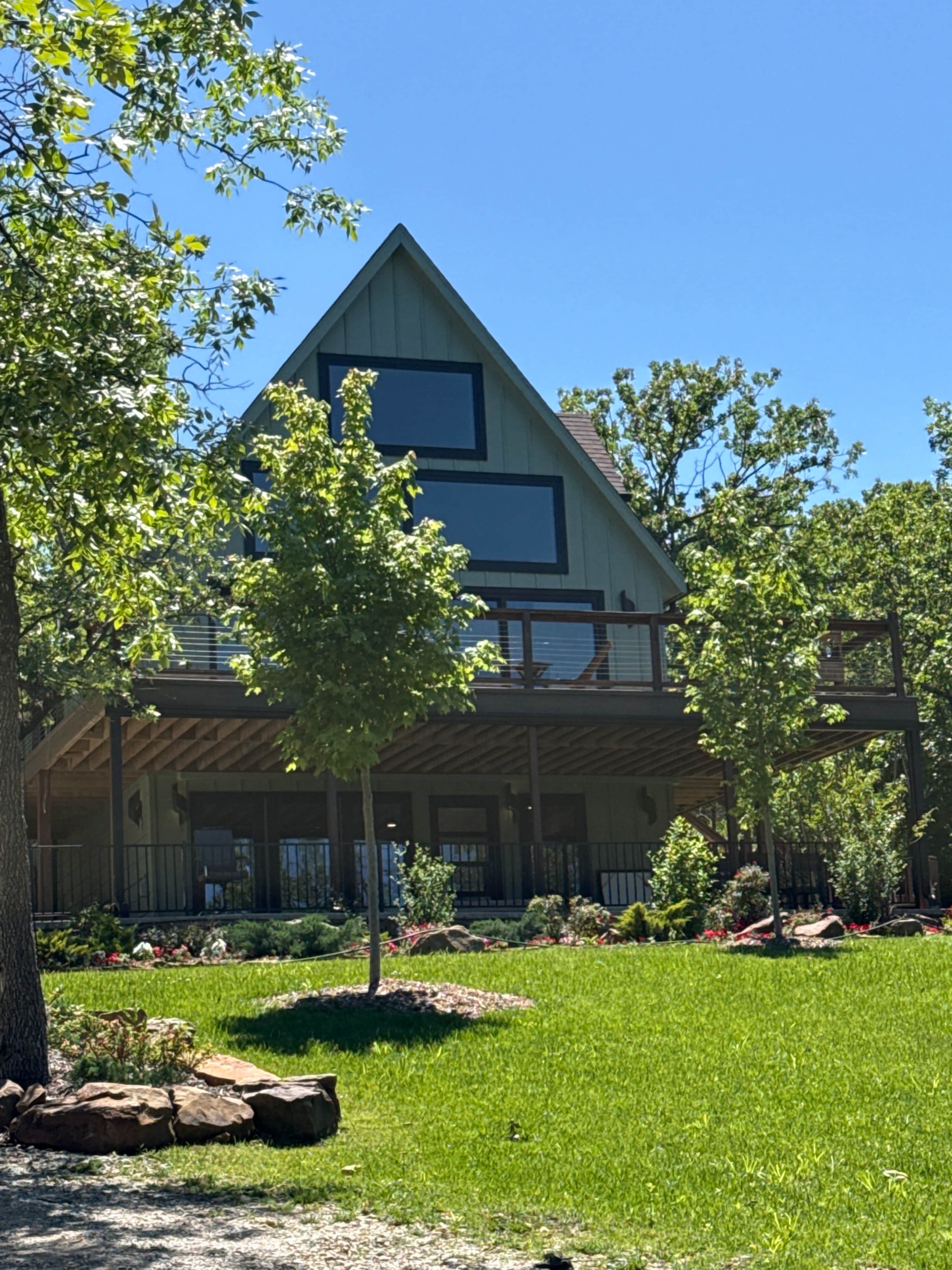 A large, triangular-roofed house with a spacious deck sits amidst green grass and landscaped gardens.