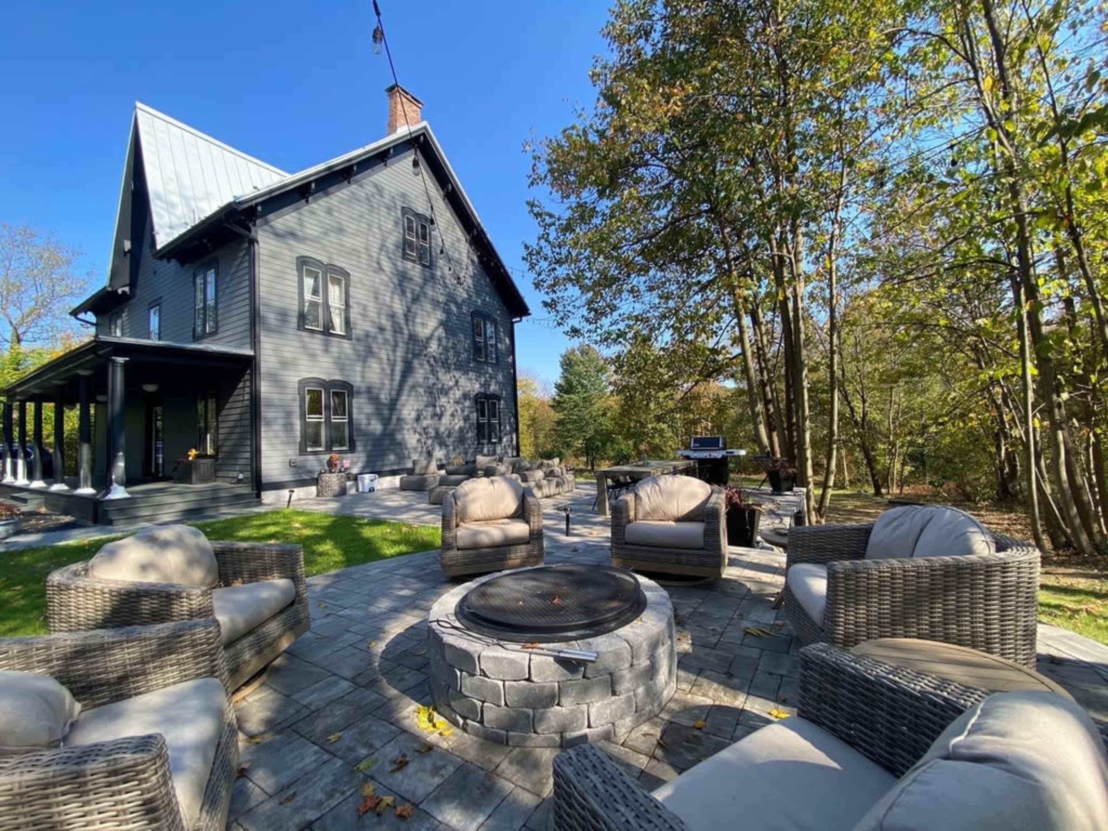 A gray house with a steep roof is surrounded by trees, featuring a stone patio with seating and a fire pit.