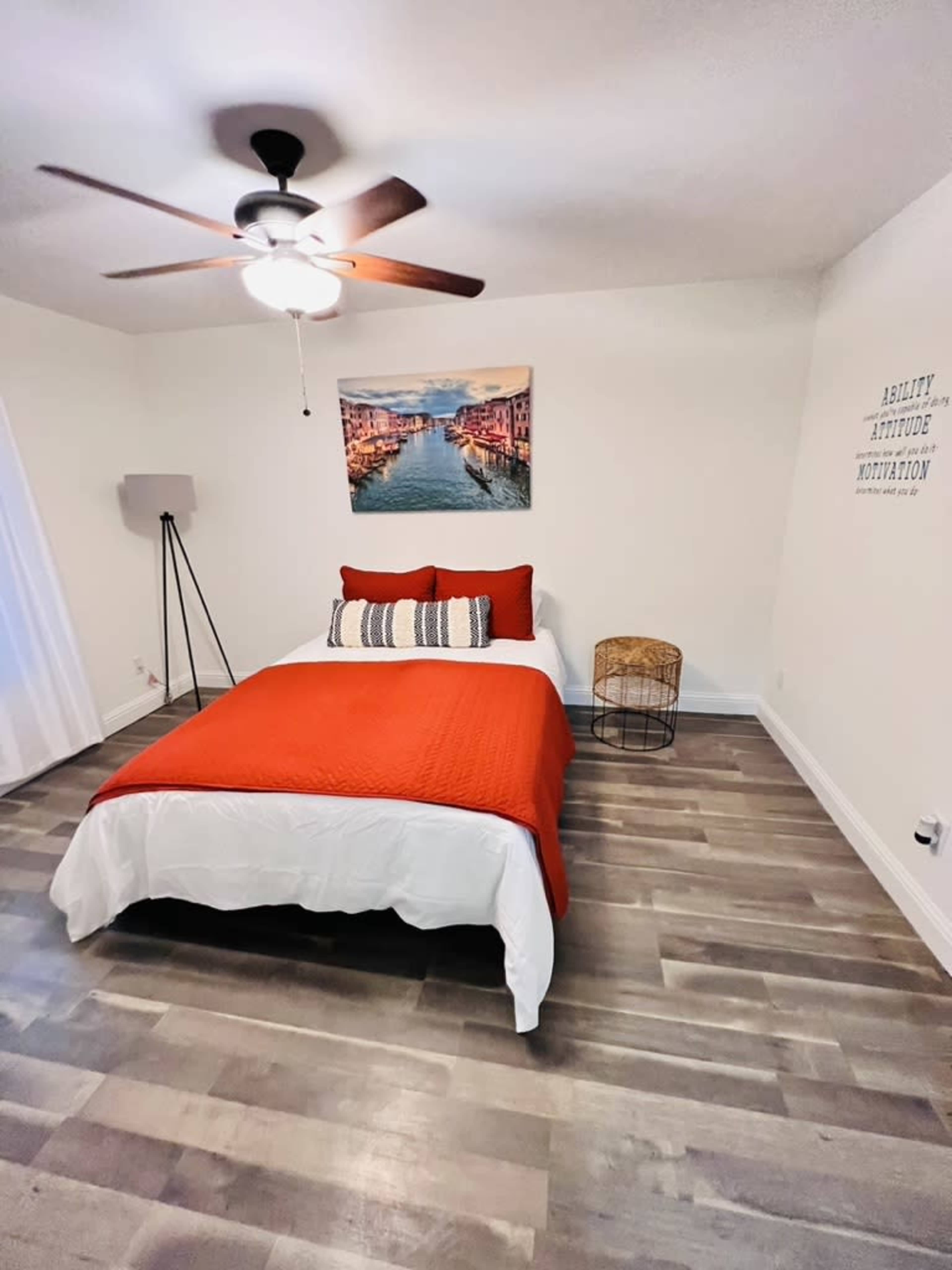 A clean and minimalistic bedroom with a bed covered in an orange quilt, a ceiling fan, and a small side table.