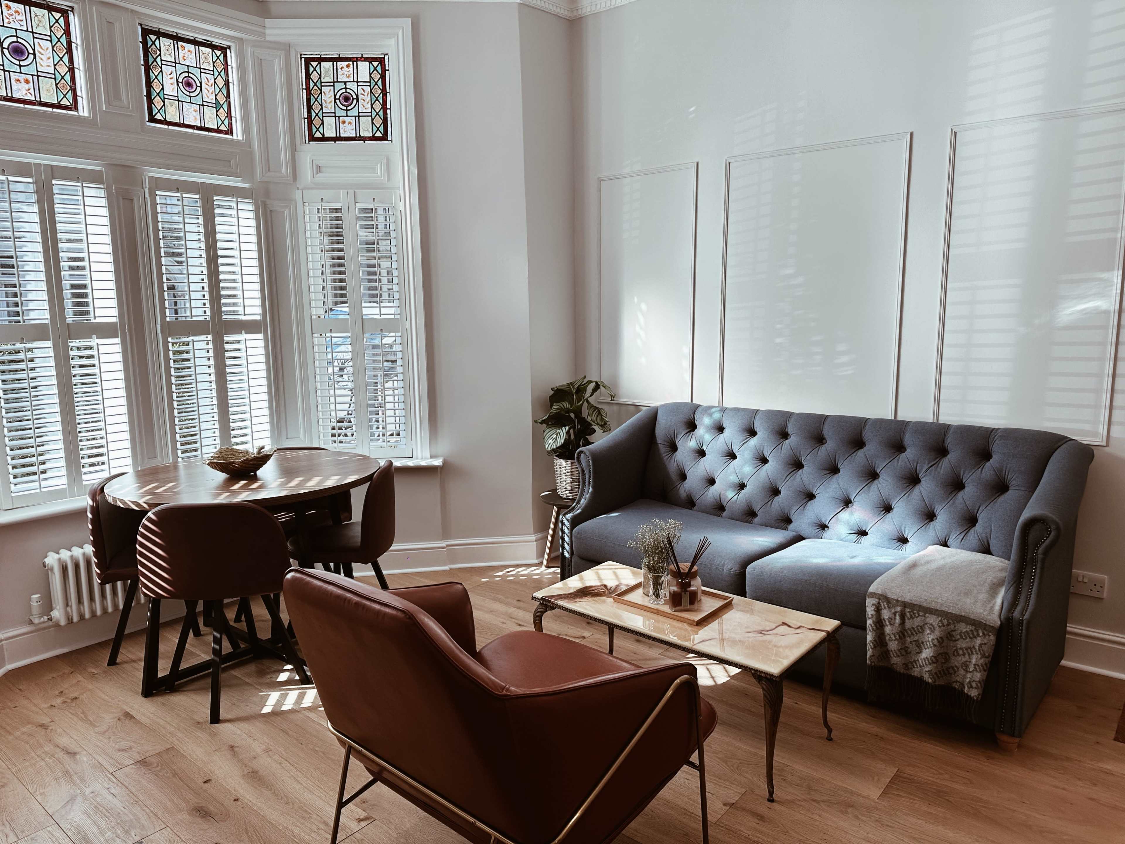 A cozy living room featuring a gray tufted sofa, a wooden coffee table, and a dining area with a round table, all illuminated by sunlight through stained glass windows.