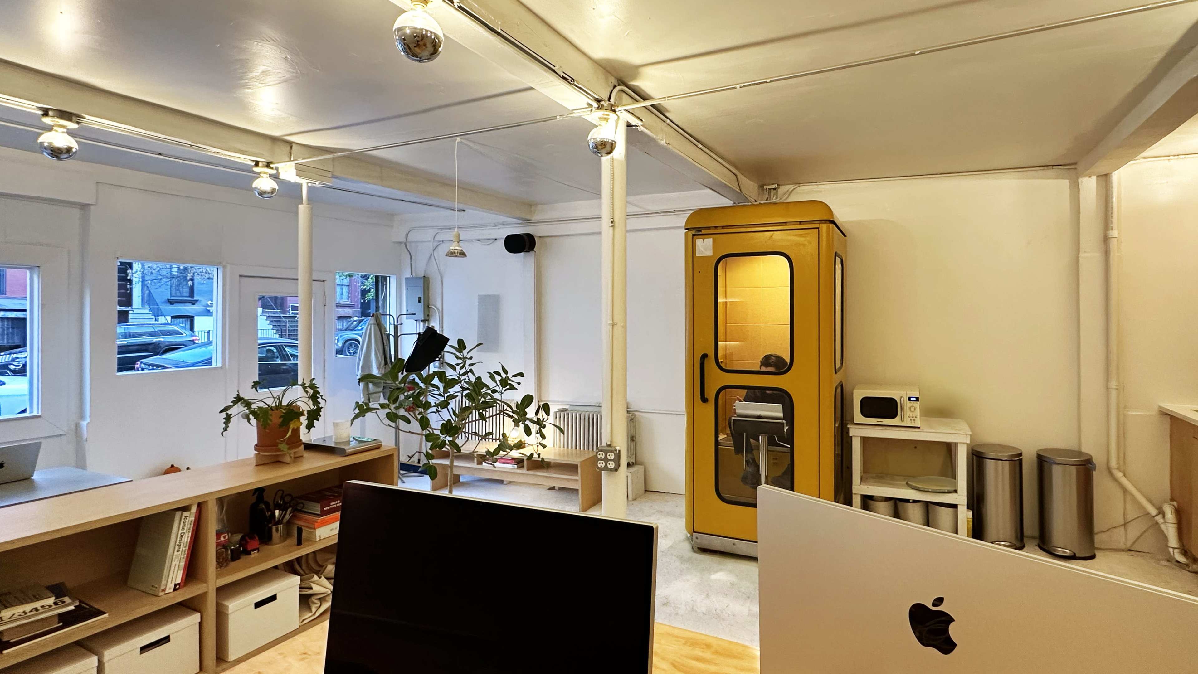 A bright, minimalist office space features a yellow phone booth, a plant, and modern workstations with Macintosh computers.