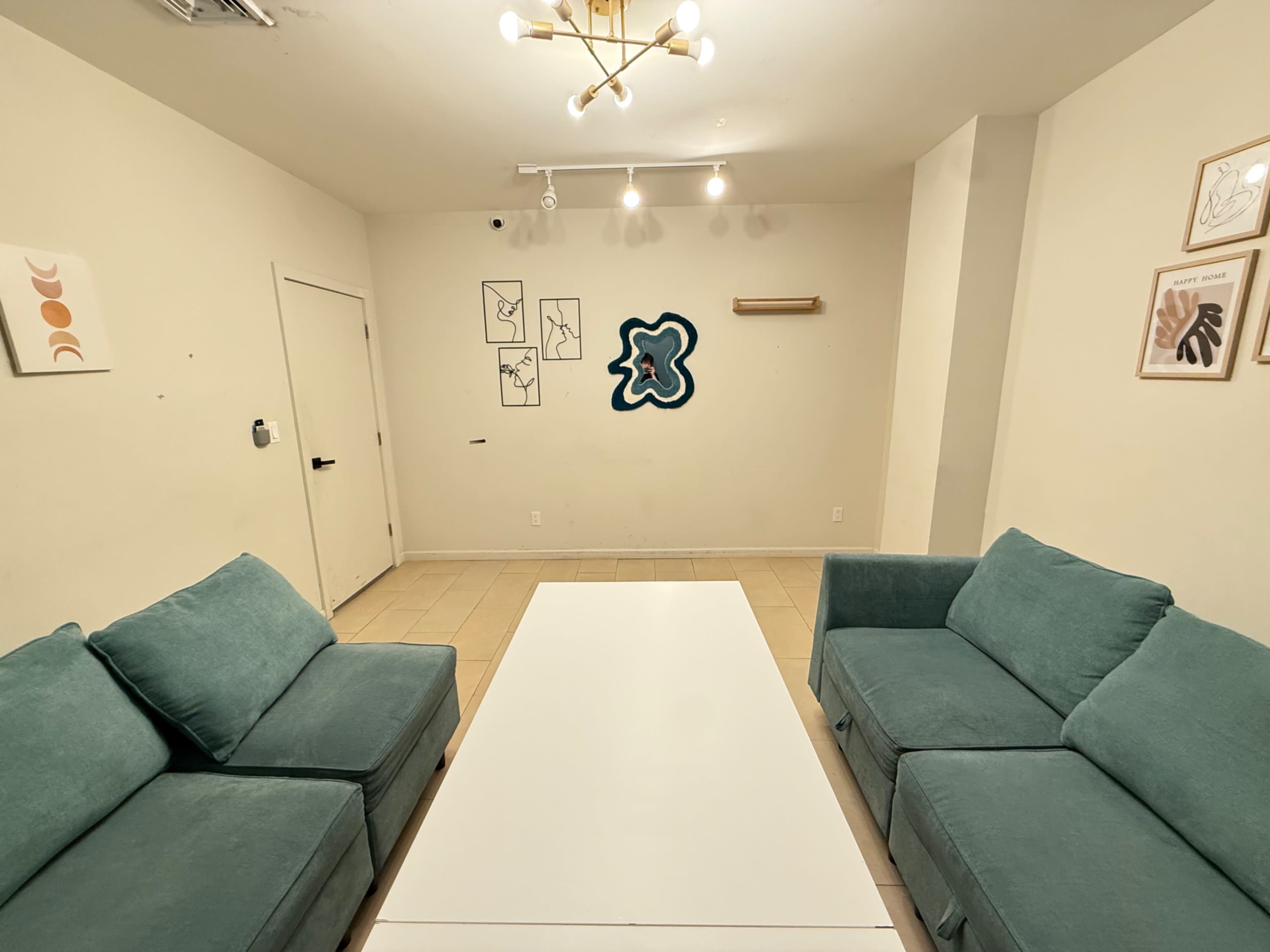 The image shows a minimalist room with two teal couches facing a white coffee table, accompanied by wall art and a door on the left.
