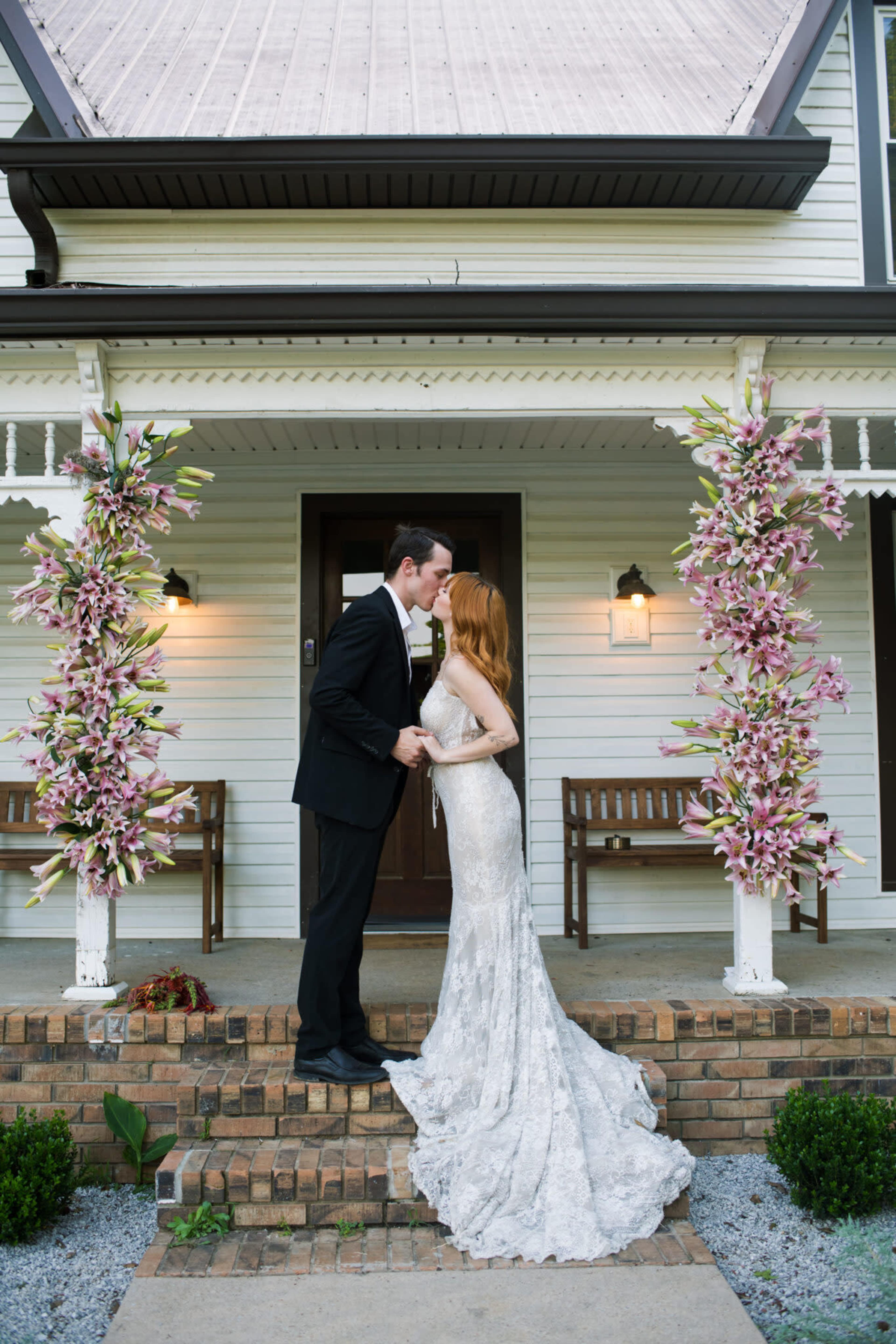 A couple in formal attire shares a kiss on the porch of a house decorated with floral arrangements.