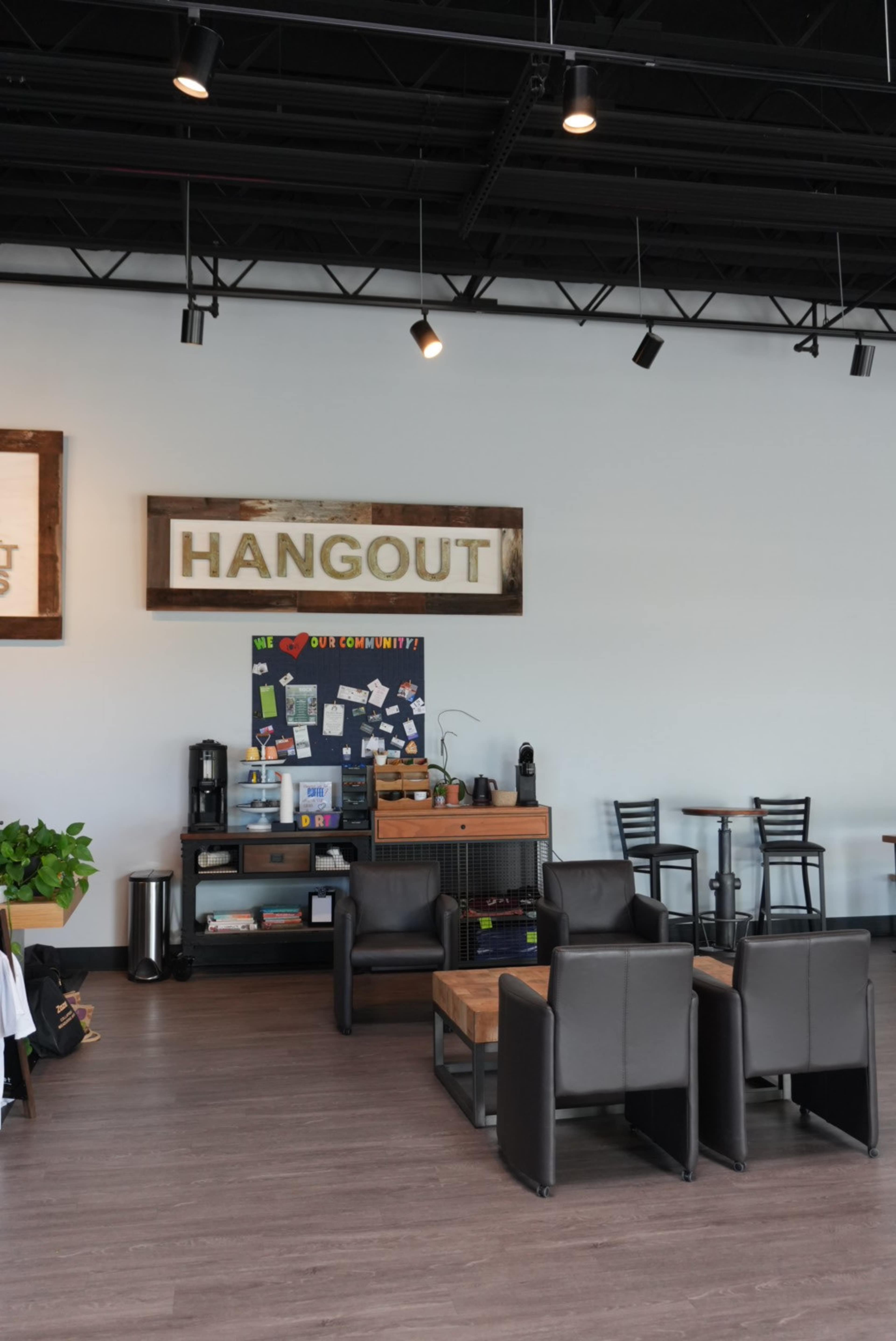 The image shows a modern cafe interior with a sign that reads "HANGOUT," featuring seating arrangements of black chairs and wooden tables, along with a coffee station and a bulletin board.